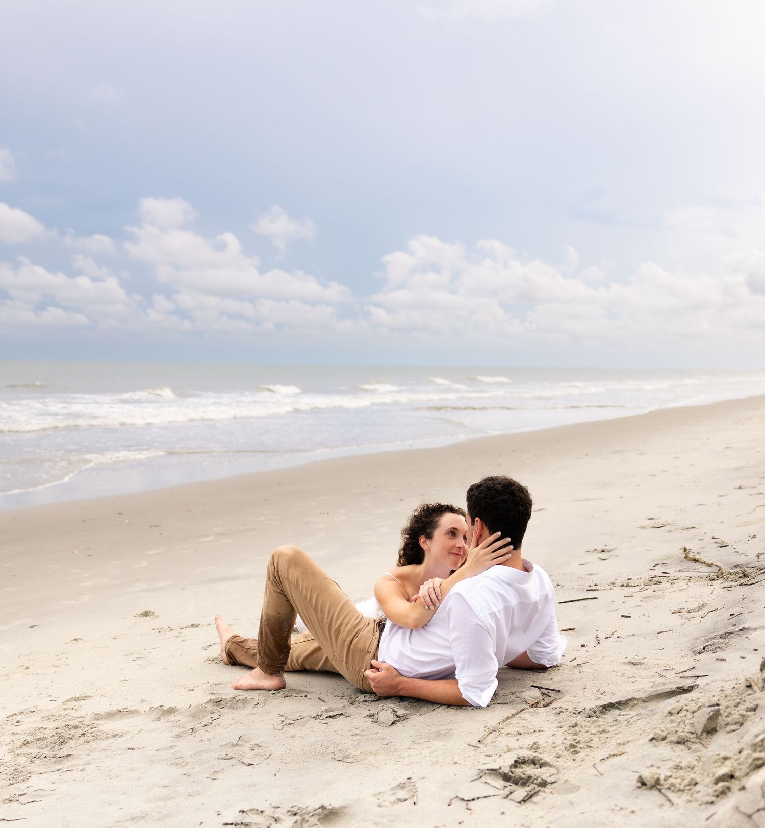 Romantic couple sitting together on a sandy beach, sharing a tender moment with ocean waves in the background.