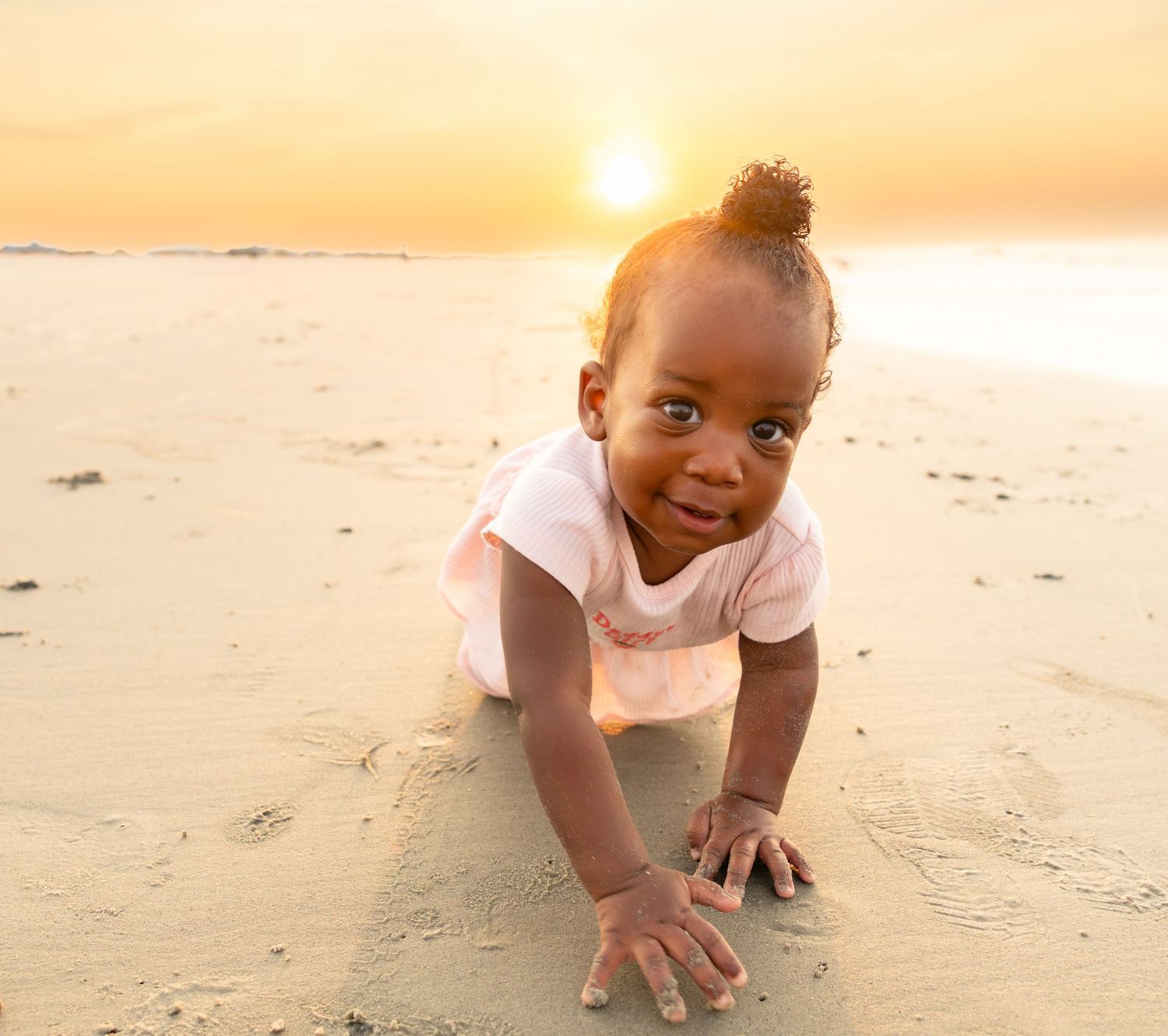 A cute toddler girl crawls on sandy beach at golden sunset, looking at camera with bright curious eyes.