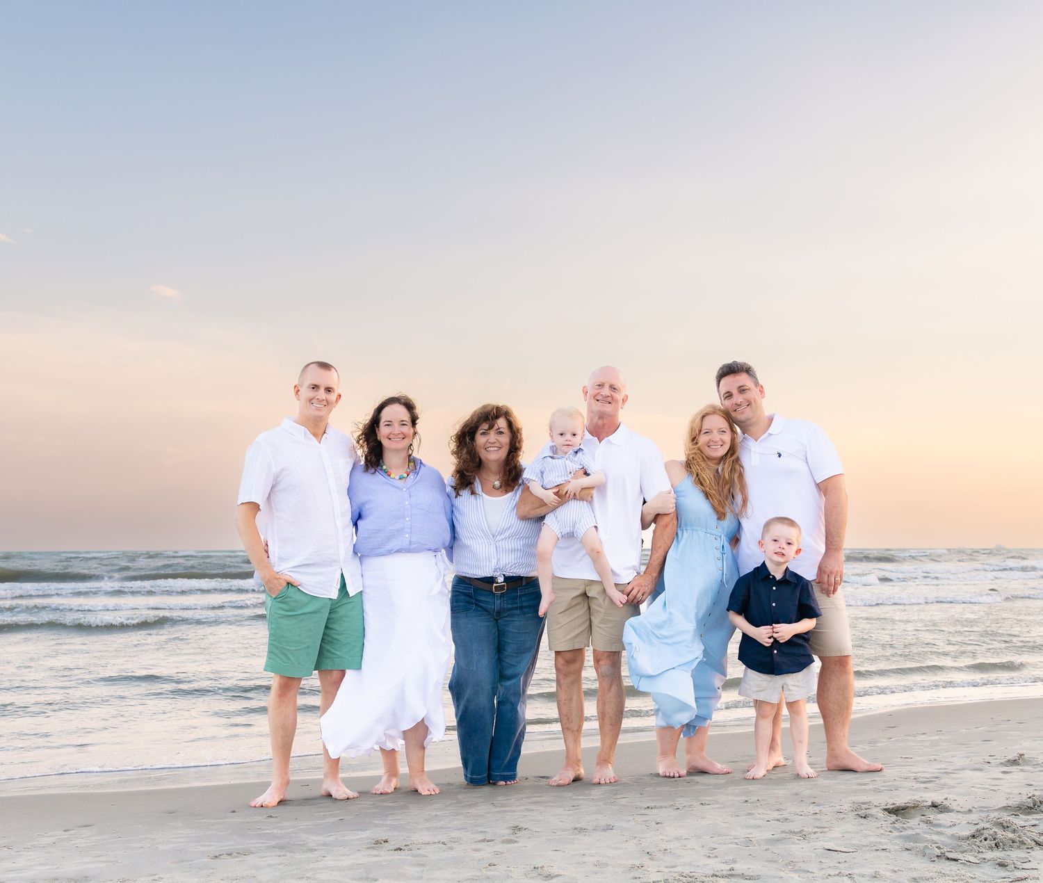 Multi-generational family posing together on a sandy beach at sunset, dressed in coordinating white and blue outfits.