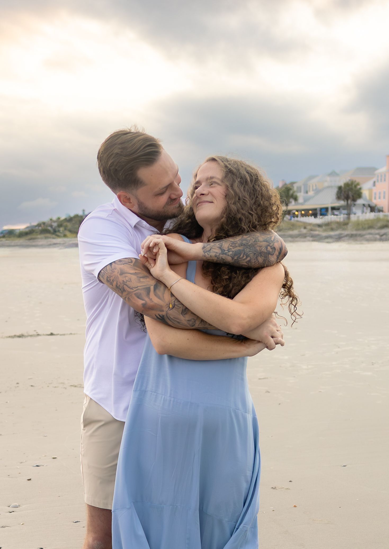 Couple embracing on a beach at sunset, woman in blue dress and man with tattoos, smiling at each other with cloudy sky.