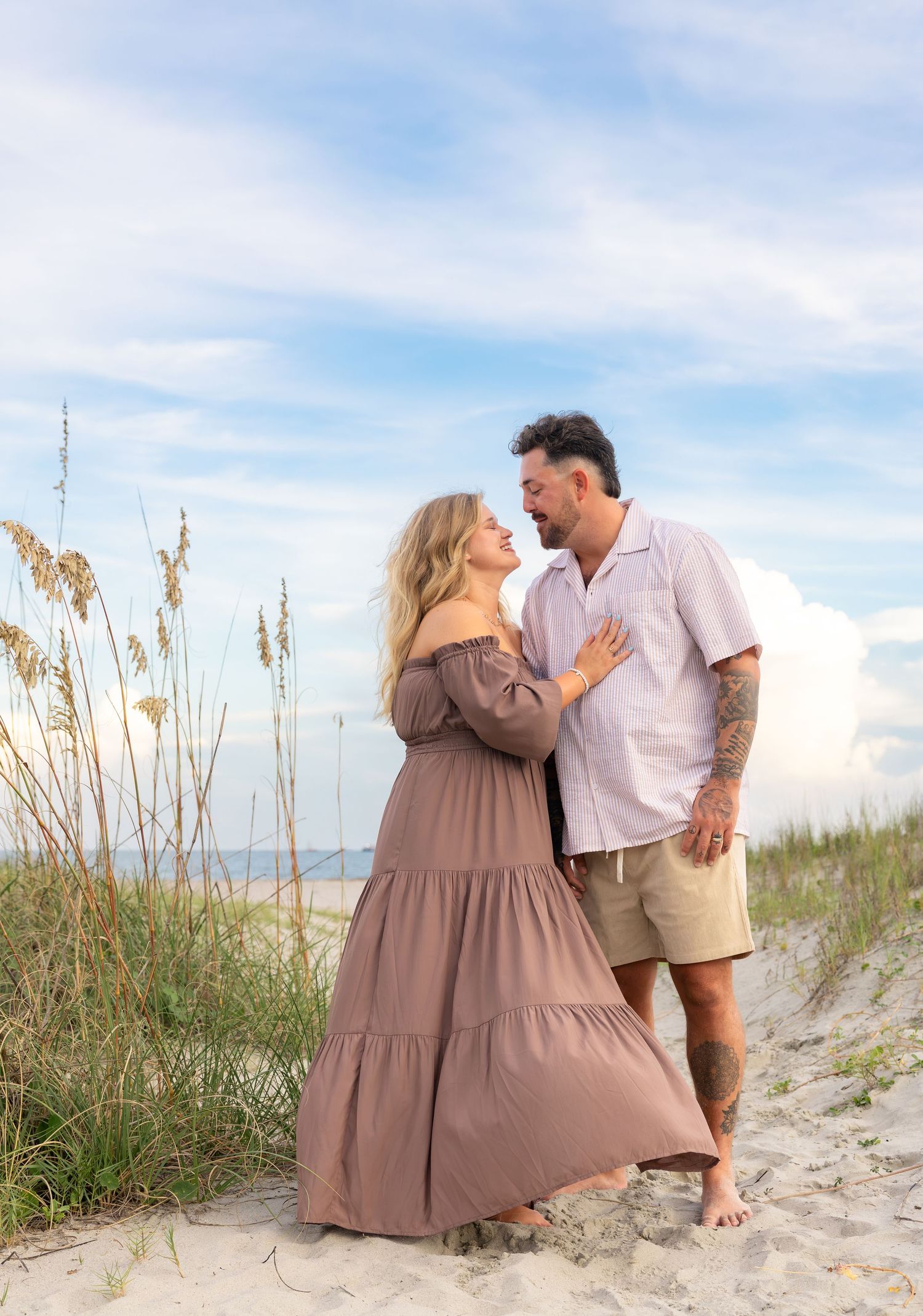 Couple embracing and smiling on a sandy beach dune with sea oats, she in a flowing mauve maxi dress, he in a linen shirt and shorts.
