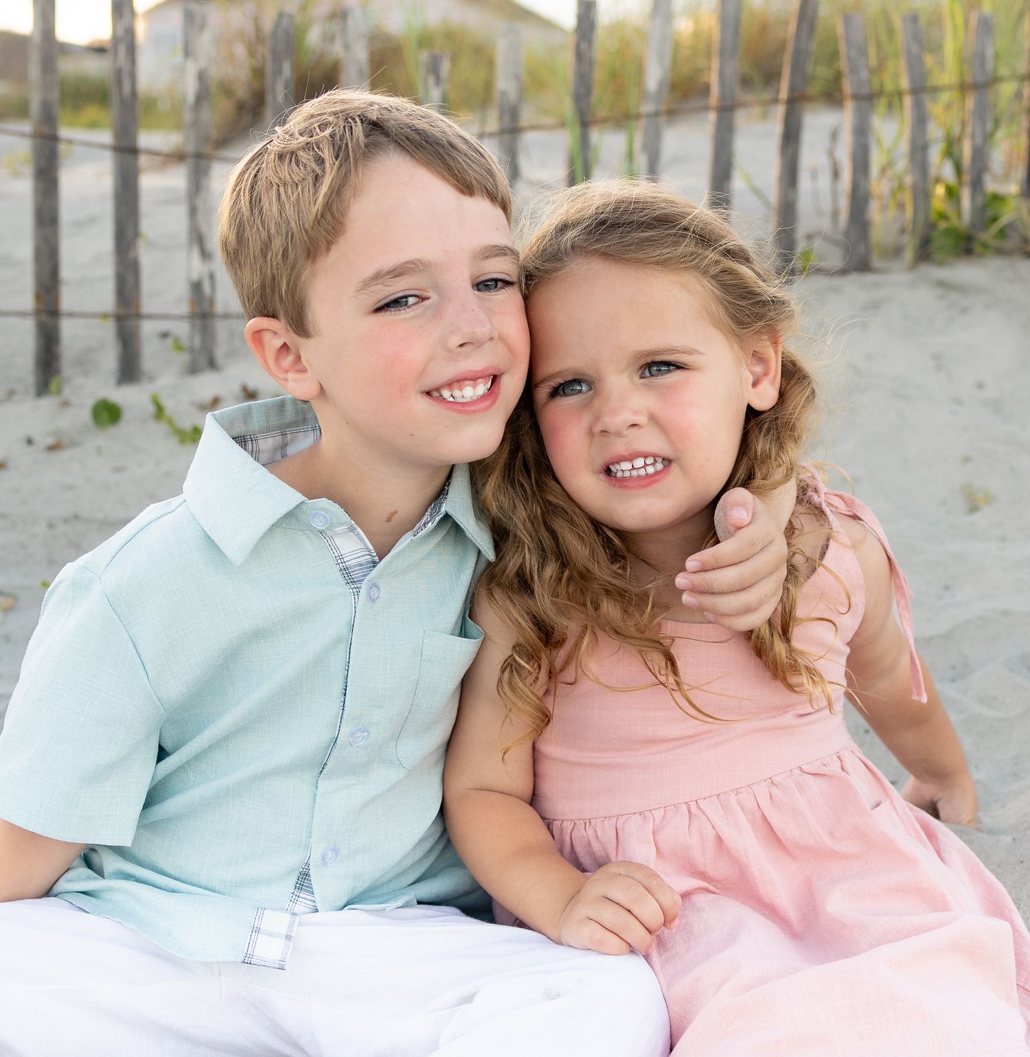 Two smiling children, a boy in light blue shirt and girl in pink dress, pose together on a beach with sand dunes and fence.