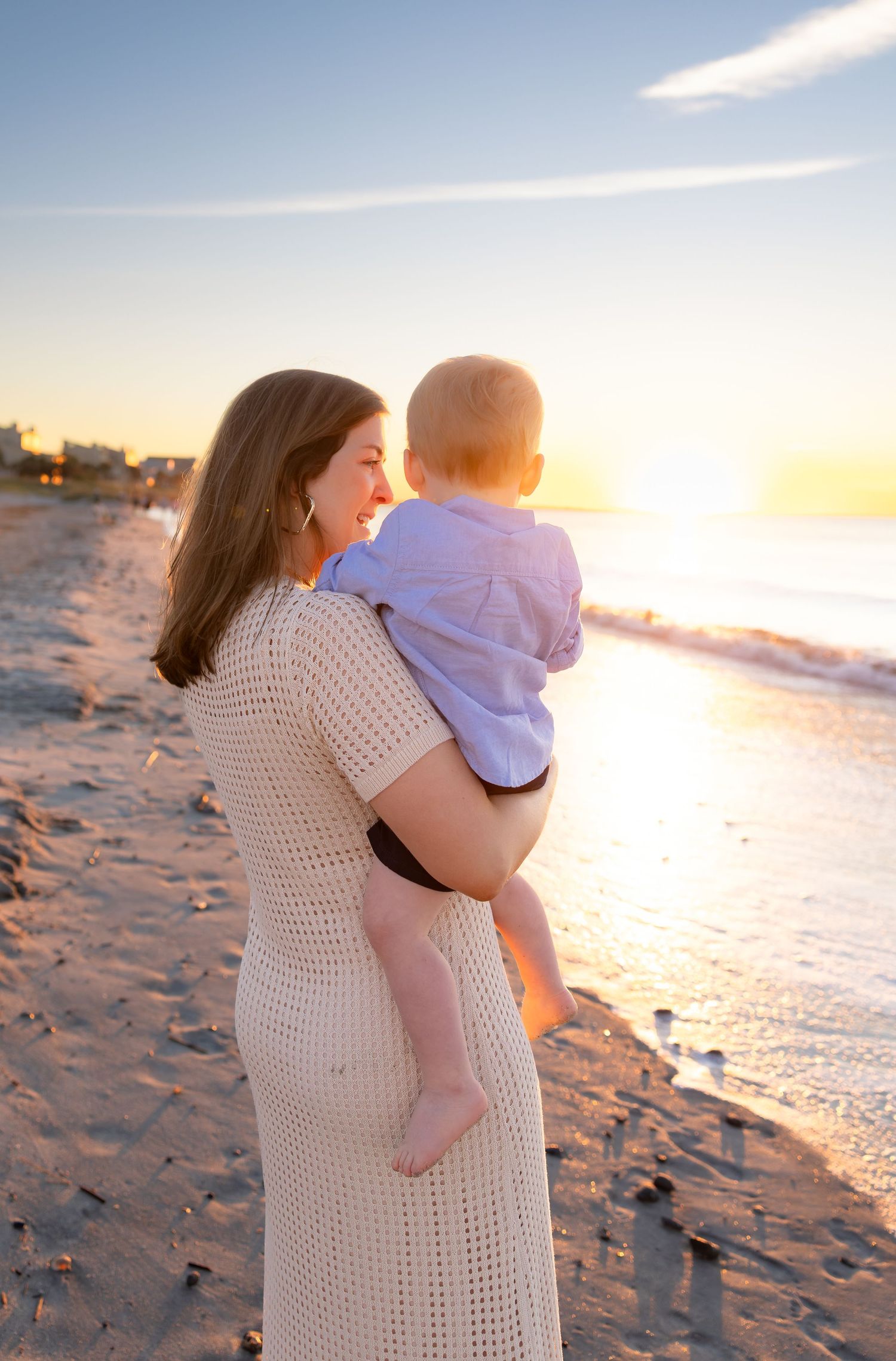 Woman in a cream knit dress holding a toddler in a lavender shirt on a sandy beach at golden sunset beside gentle ocean waves.