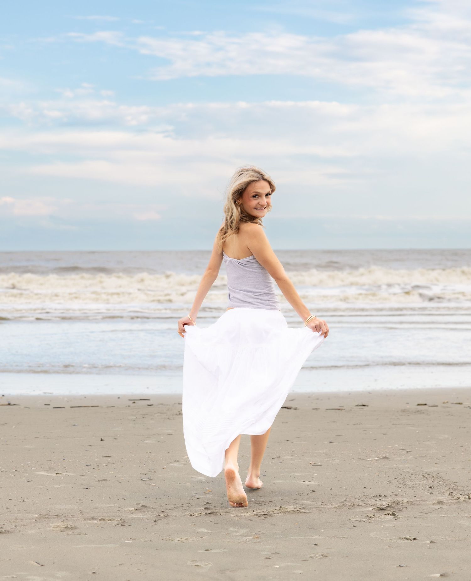 Smiling young woman in a gray top and flowing white skirt walks barefoot on a sandy beach with ocean waves and a cloudy blue sky behind her.