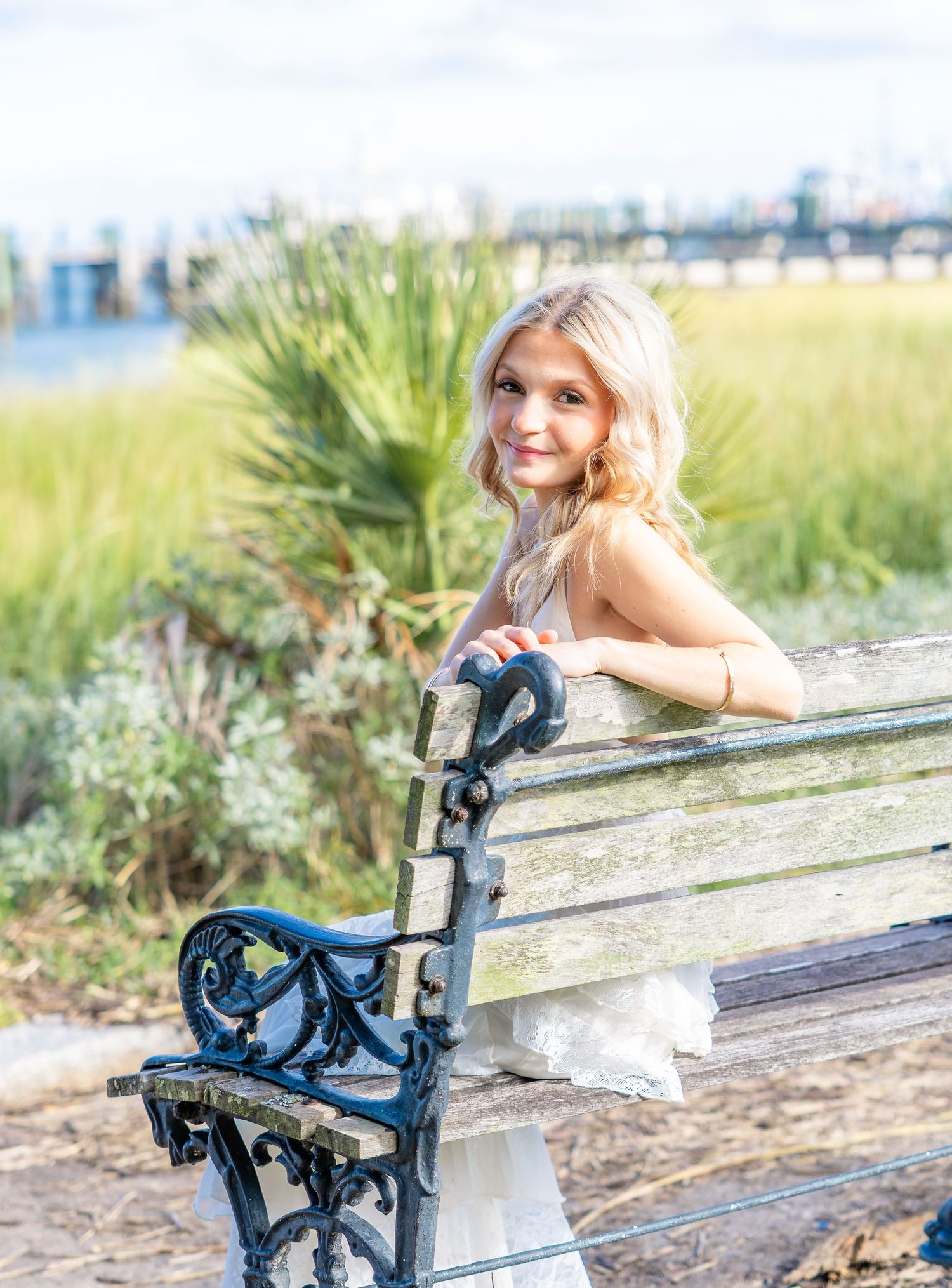 Smiling young woman with long blonde hair in a white dress sits on a weathered wooden bench with ornate iron armrests near a coastal marsh.