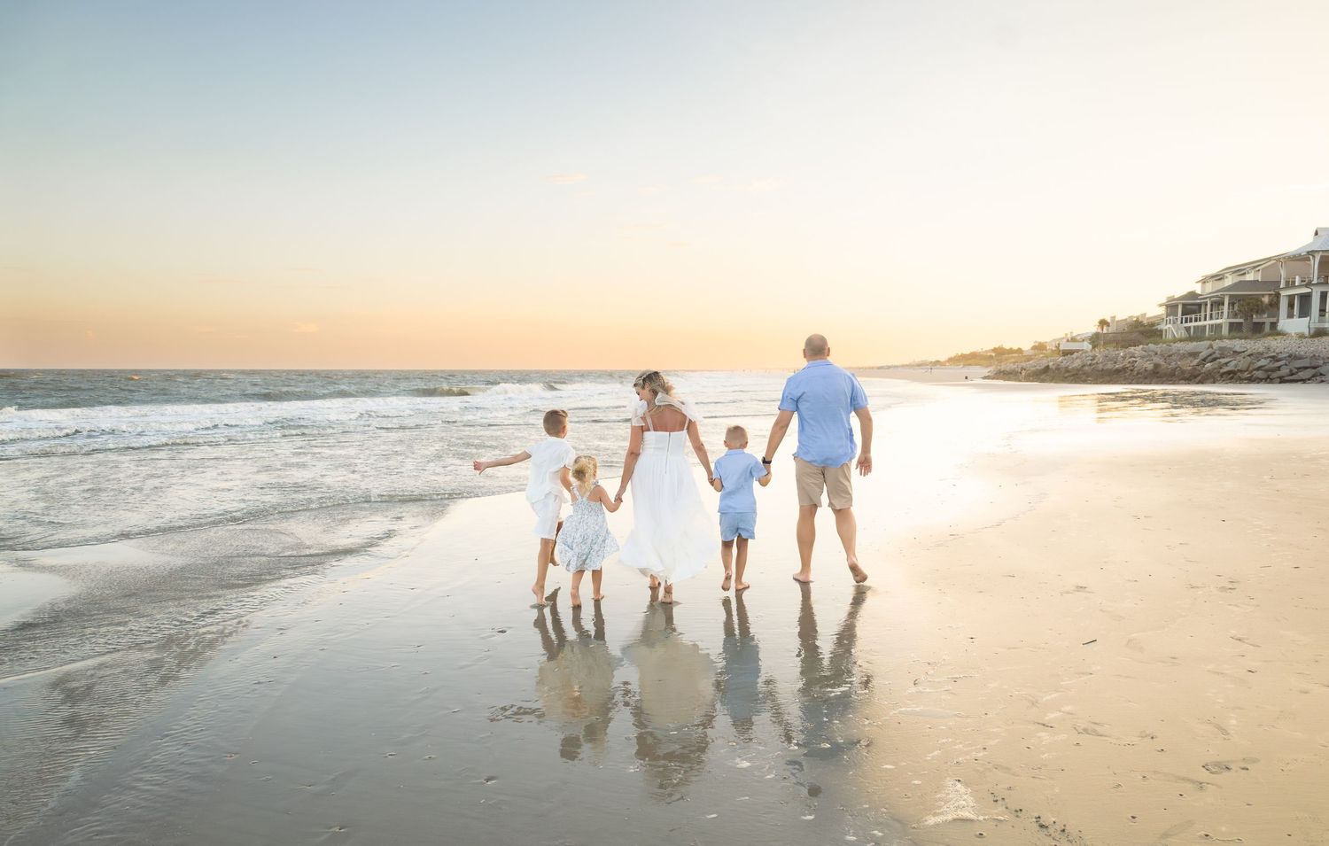 Family of four holding hands walking along a sandy beach at sunset with gentle waves and coastal homes in the background.