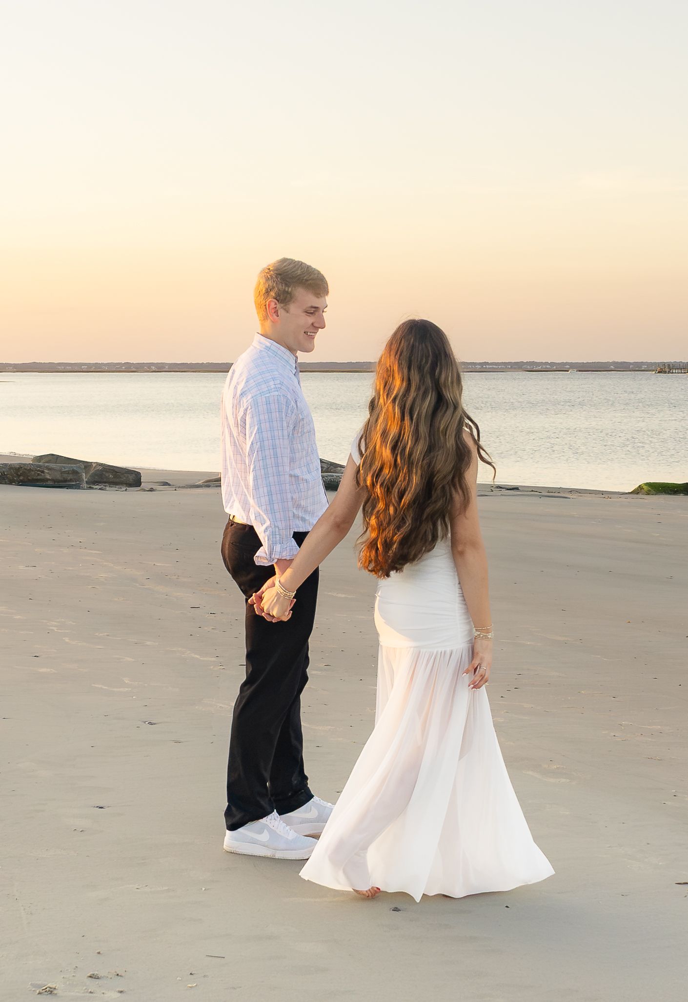 Couple holding hands on a sandy beach at sunset, woman in white dress with long wavy hair, man in plaid shirt.