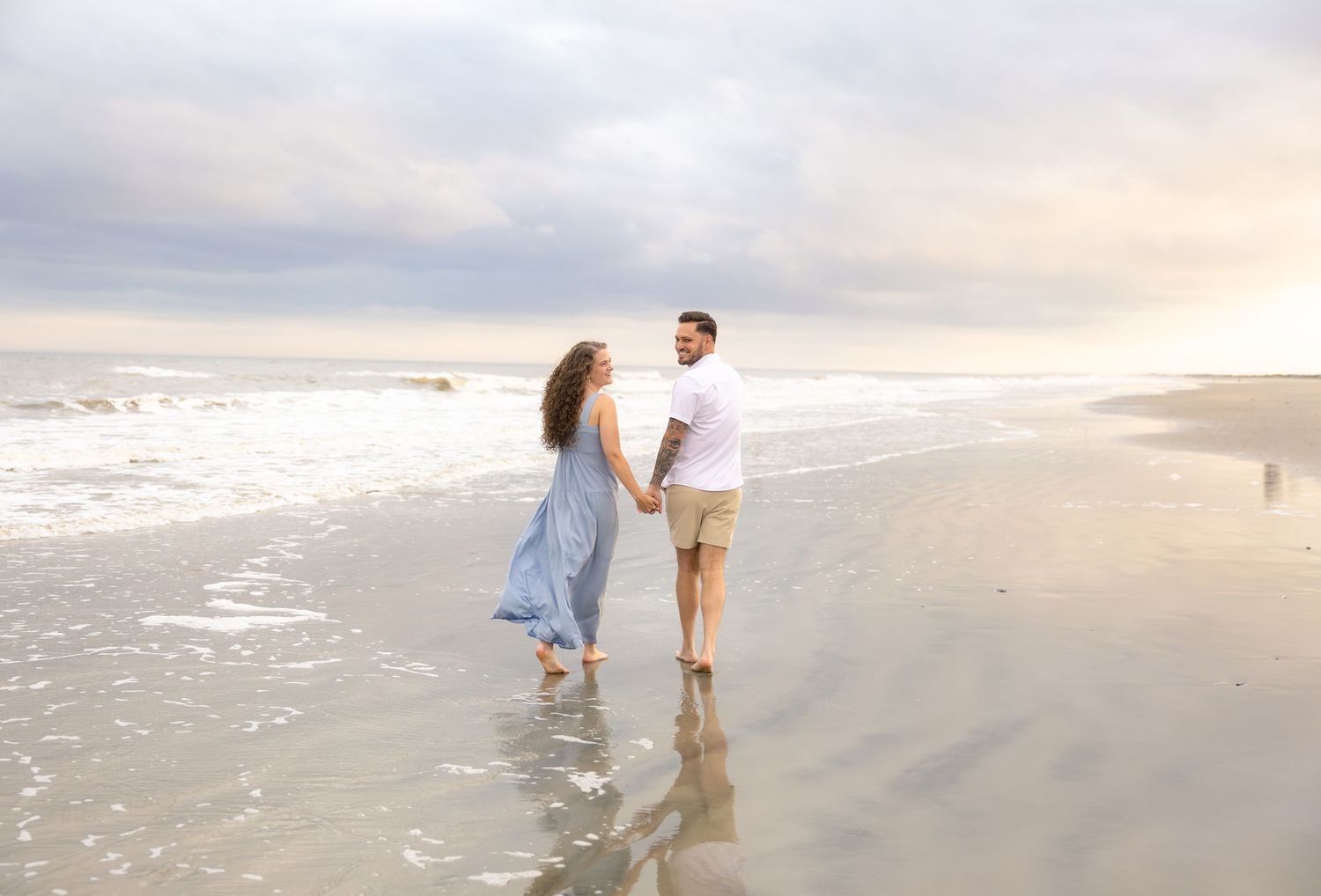 A couple holds hands while walking barefoot along a serene beach with gentle waves and a cloudy sky.