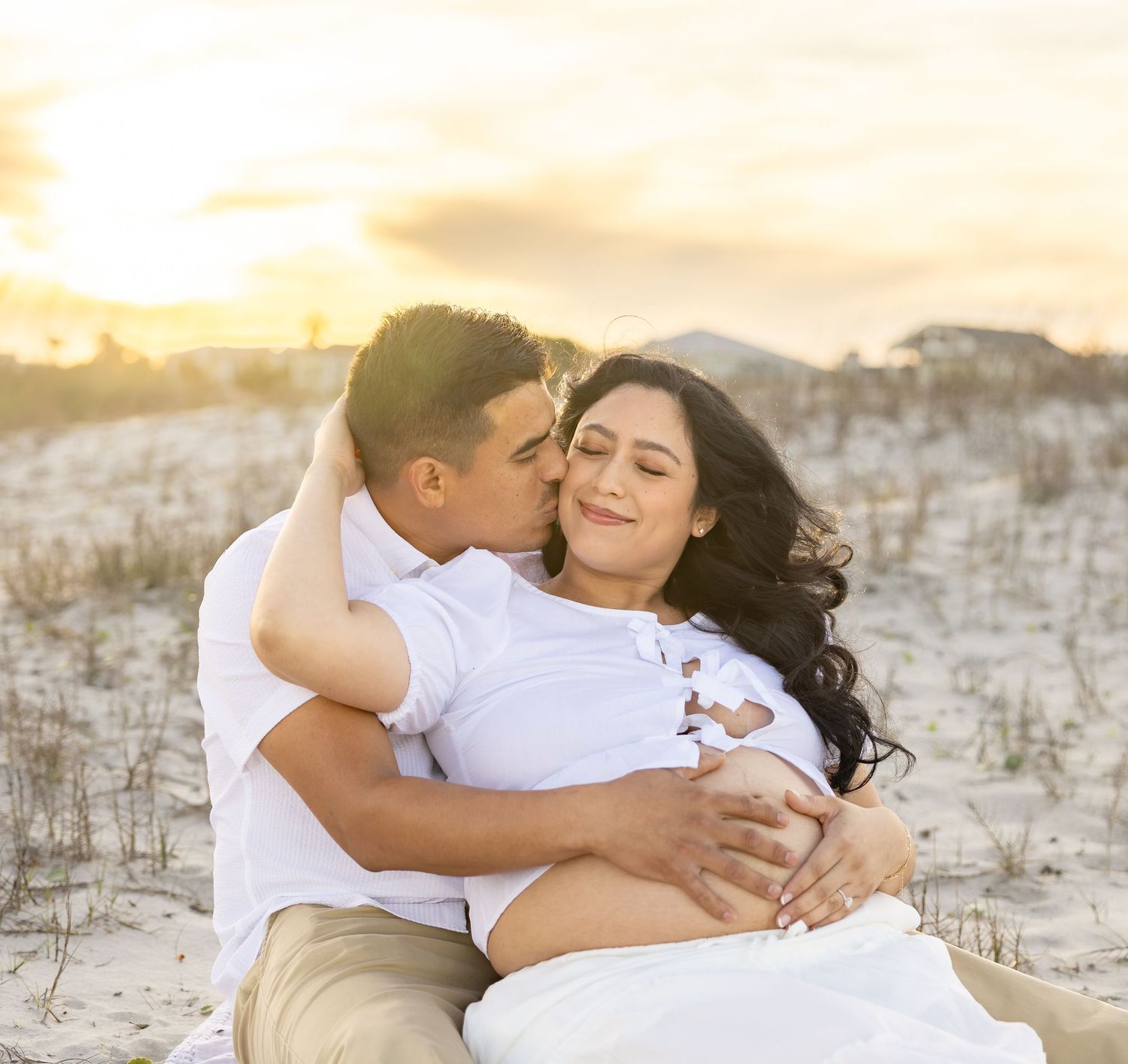 Couple sharing a romantic embrace on a sandy beach at sunset, woman in white dress with baby bump.