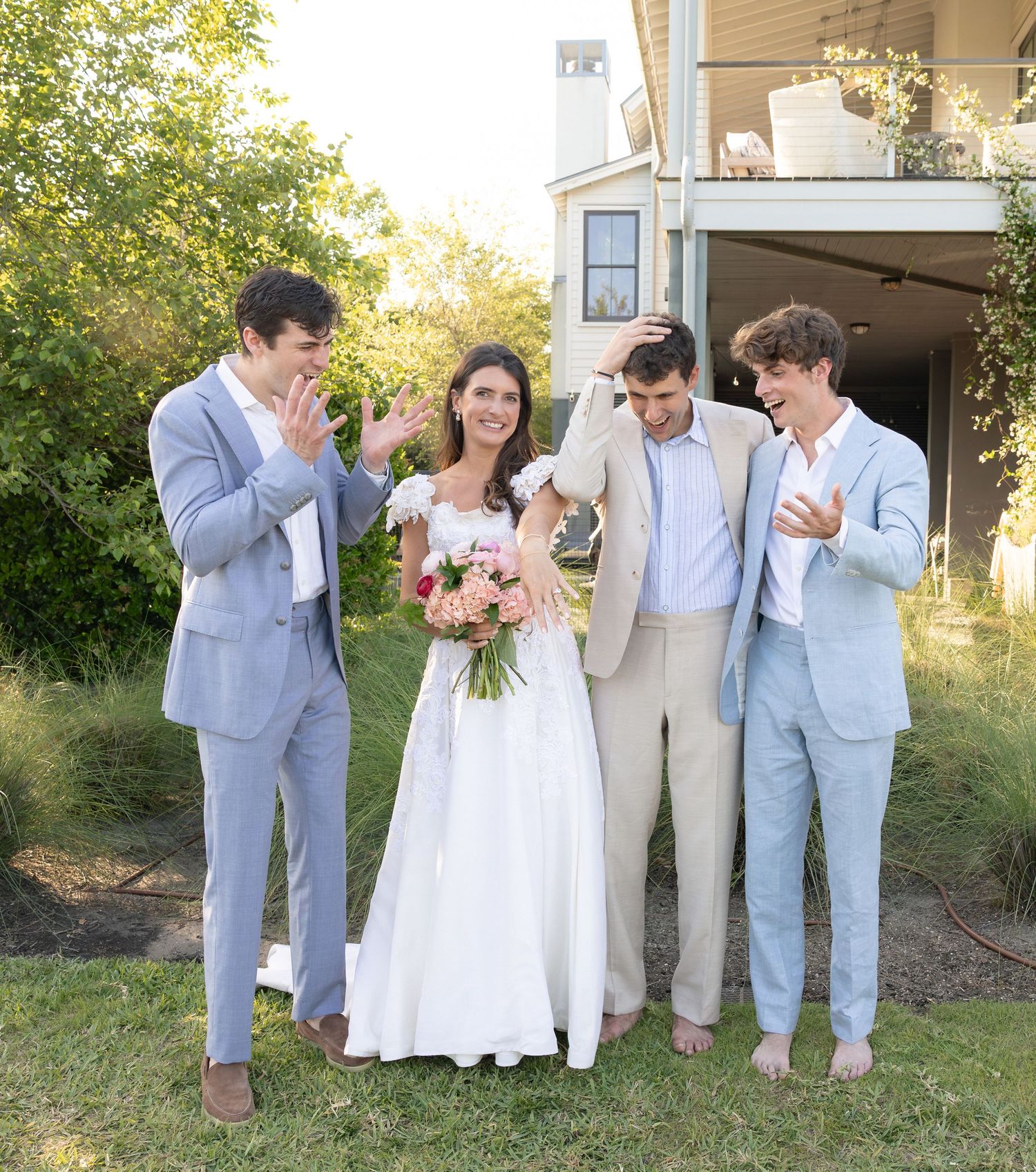 Bride in white gown holds pink bouquet surrounded by three men in suits celebrating outdoors at a backyard wedding.