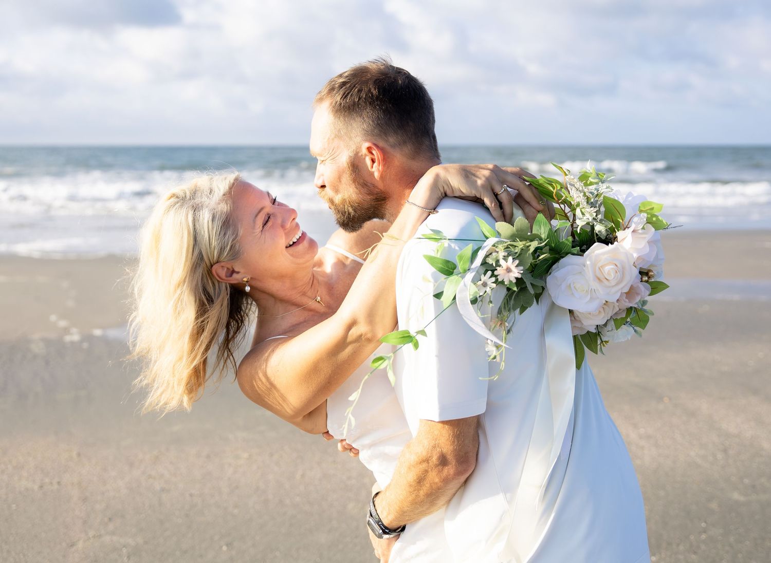 Couple sharing a romantic beach wedding moment, groom lifting bride holding a pink and white floral bouquet by the ocean.