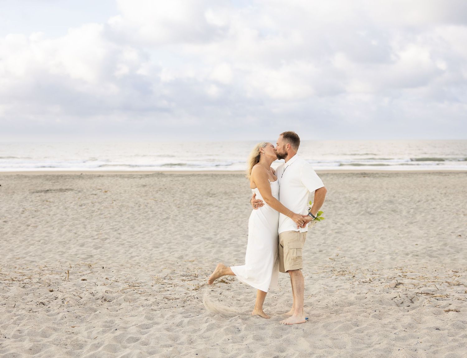 Couple kissing on sandy beach in white outfits, ocean waves and cloudy sky in background.