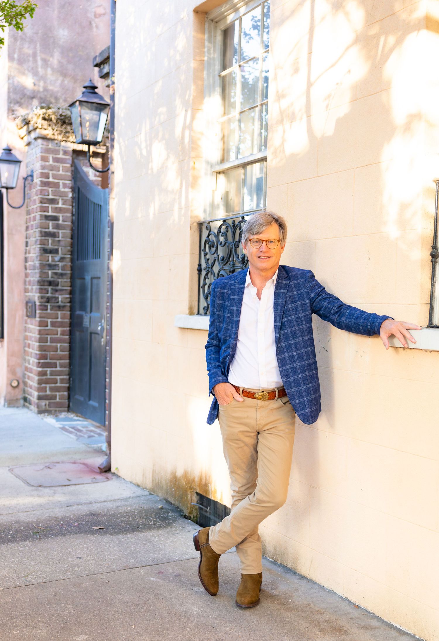Man in blue plaid blazer and khakis leaning against a light-colored wall on a charming city street.