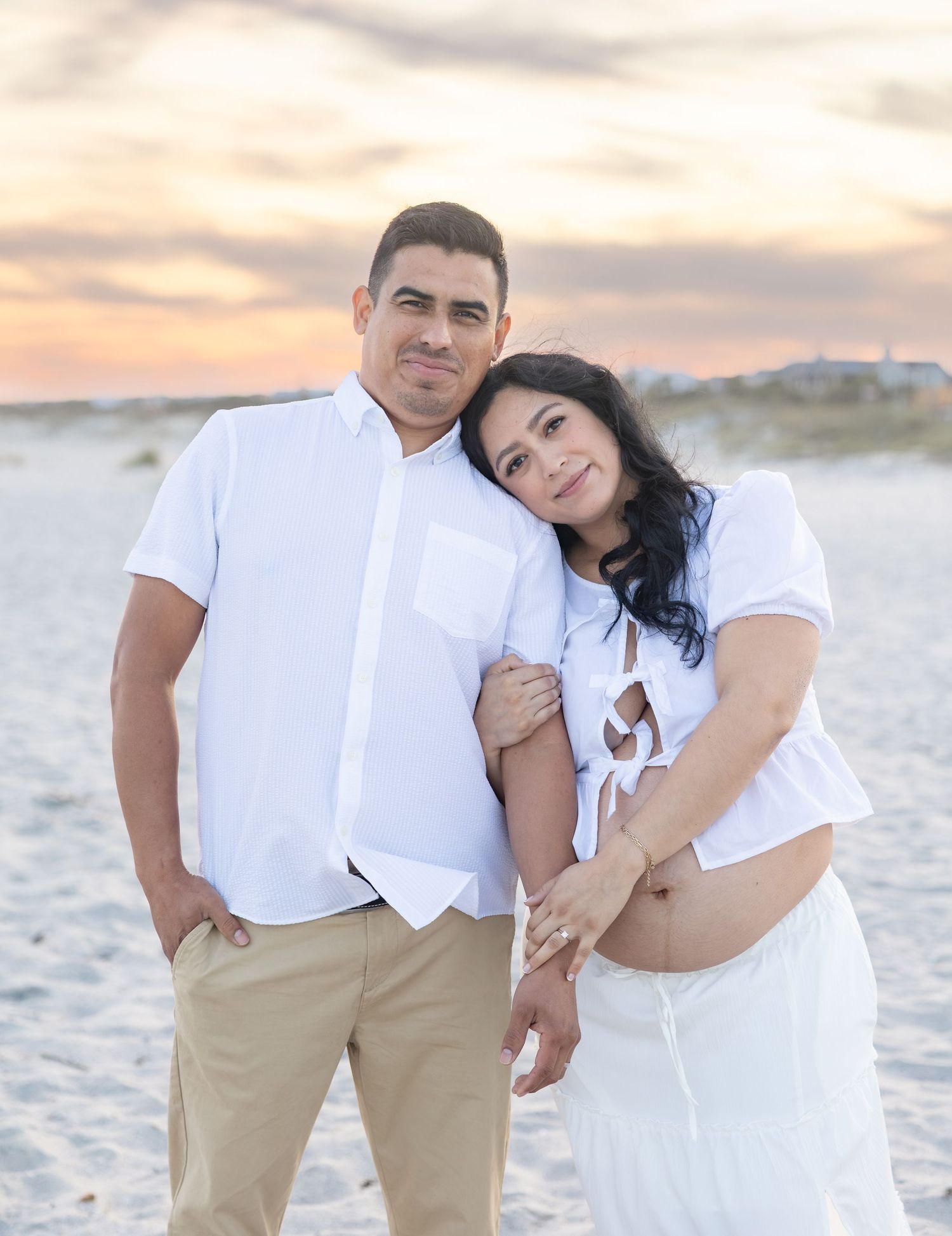 Couple in white outfits posing on sandy beach at sunset, woman showing baby bump in maternity photo session.