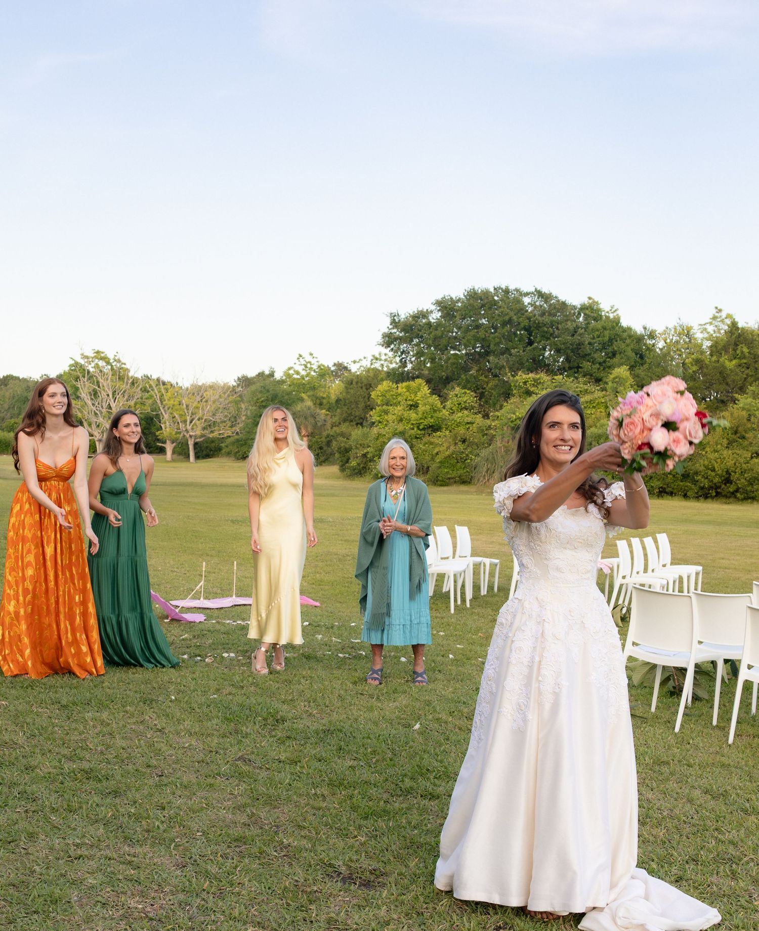 Bride in white gown tosses pink bouquet to bridesmaids in colorful dresses at outdoor wedding ceremony.