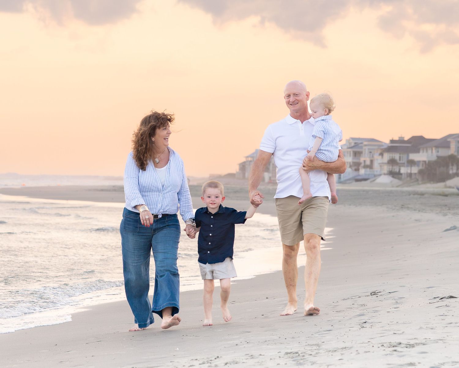Family of four walking on beach at sunset, parents holding toddler hands, father carrying baby, pink sky backdrop.