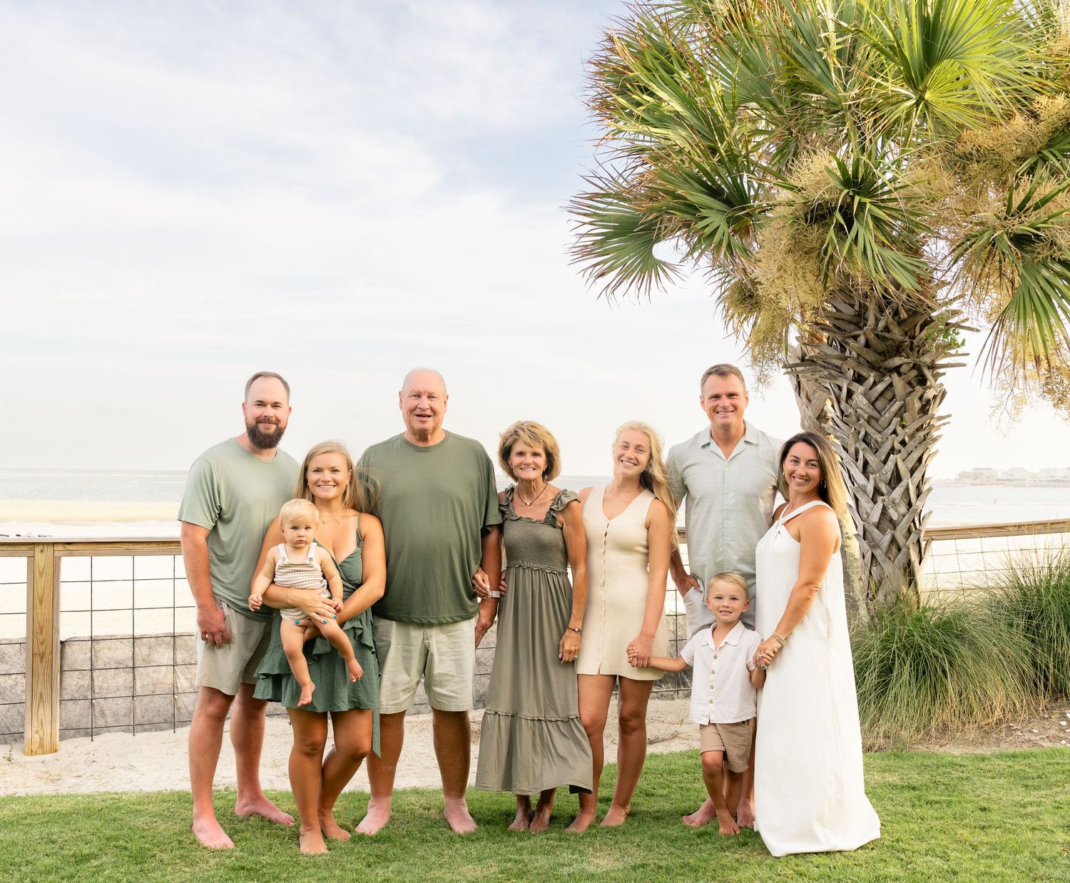Multi-generational family posing together outdoors near a beach with a palm tree and ocean views on a sunny day.