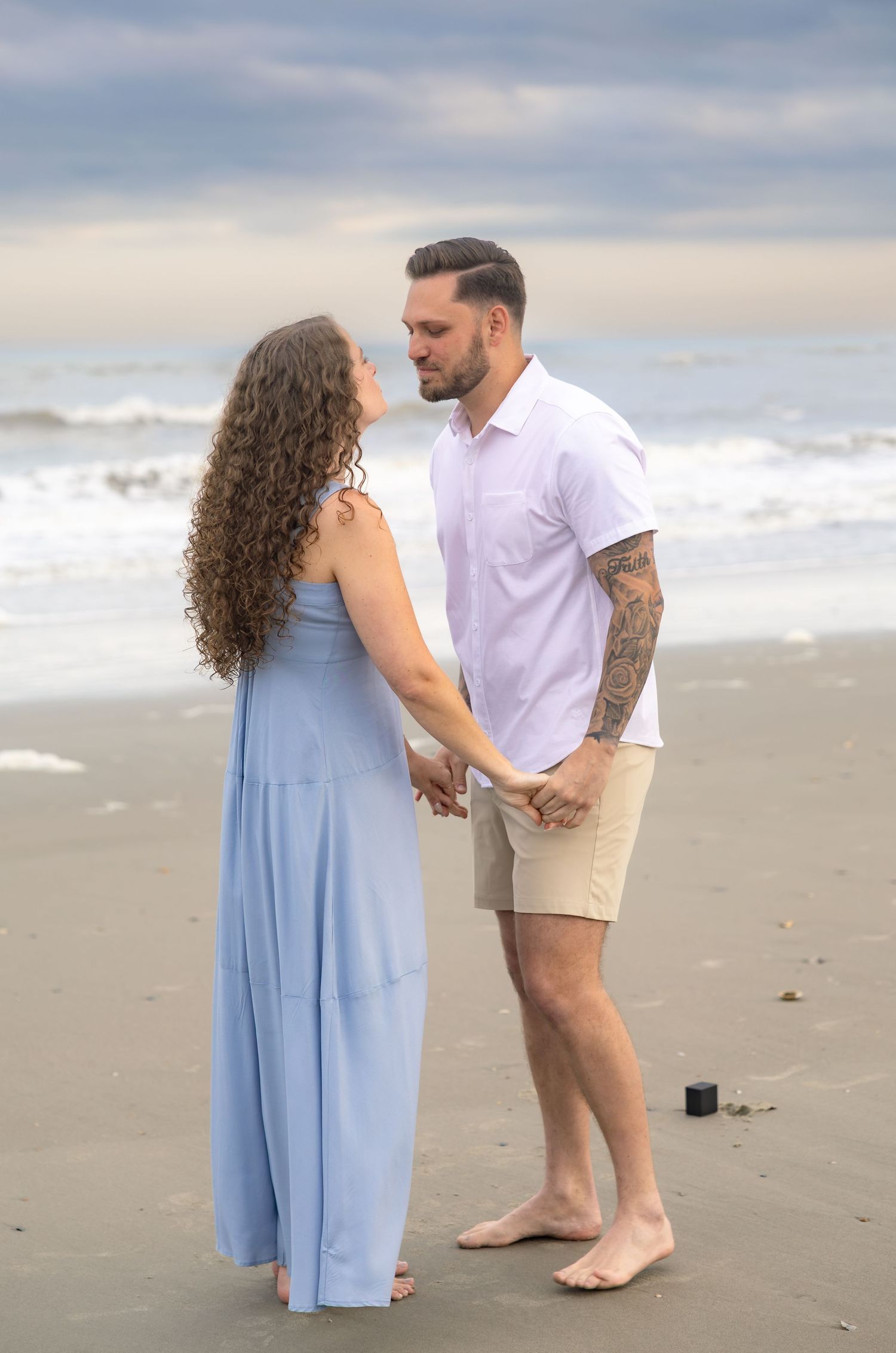 Couple holding hands on a beach at sunset, woman in blue maxi dress, man in white shirt with tattoos.