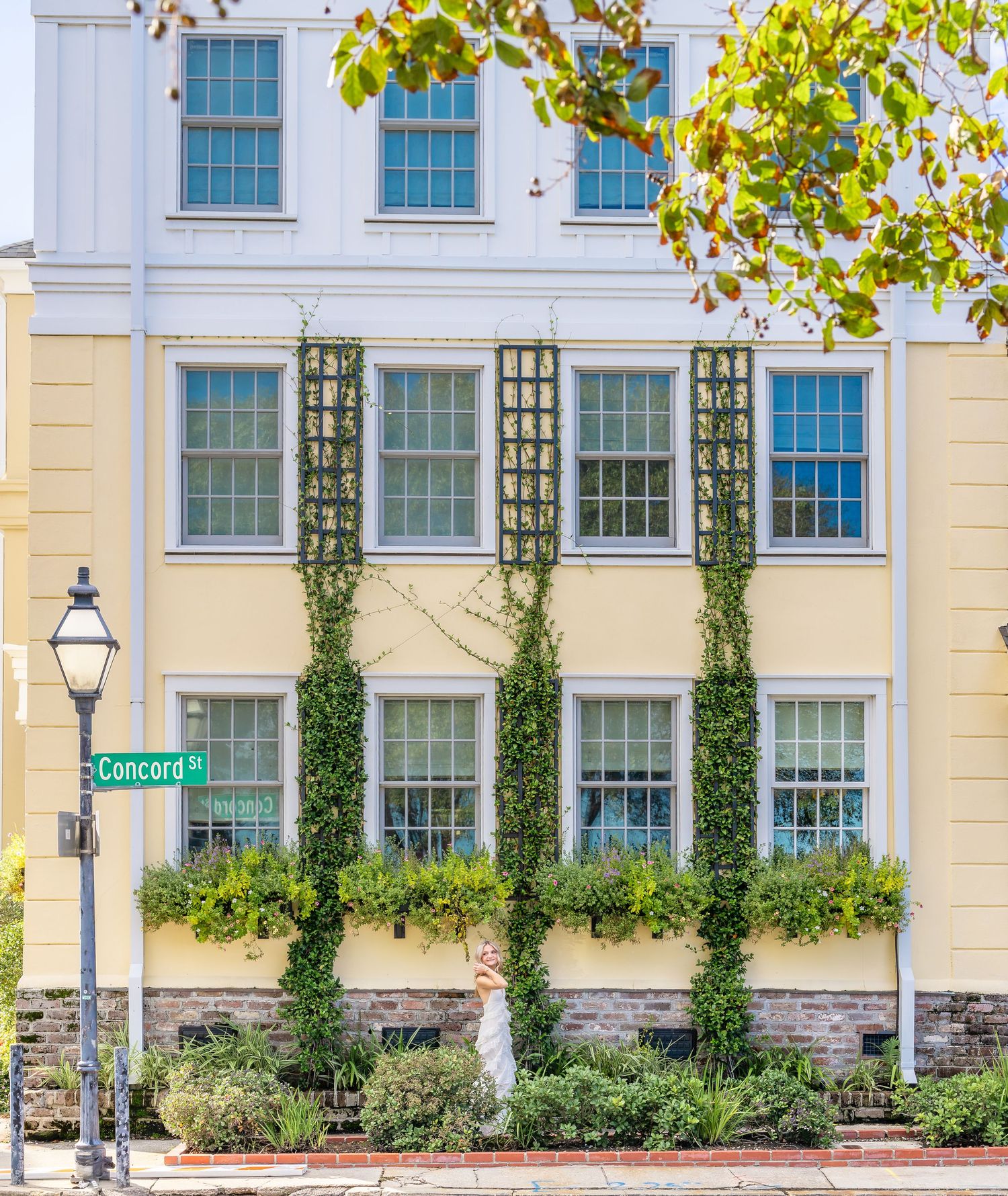 Yellow historic building with vine-covered trellises, flower-filled window boxes and a Concord St street sign beside a vintage lamppost.
