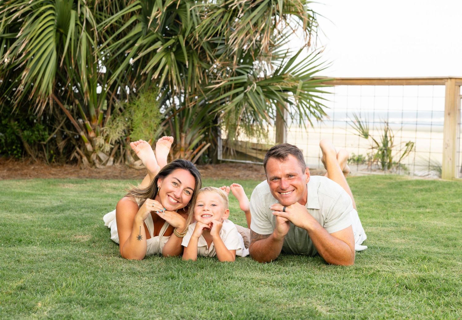 Happy family with young child lying on green grass near tropical palm trees and beach fence outdoors.