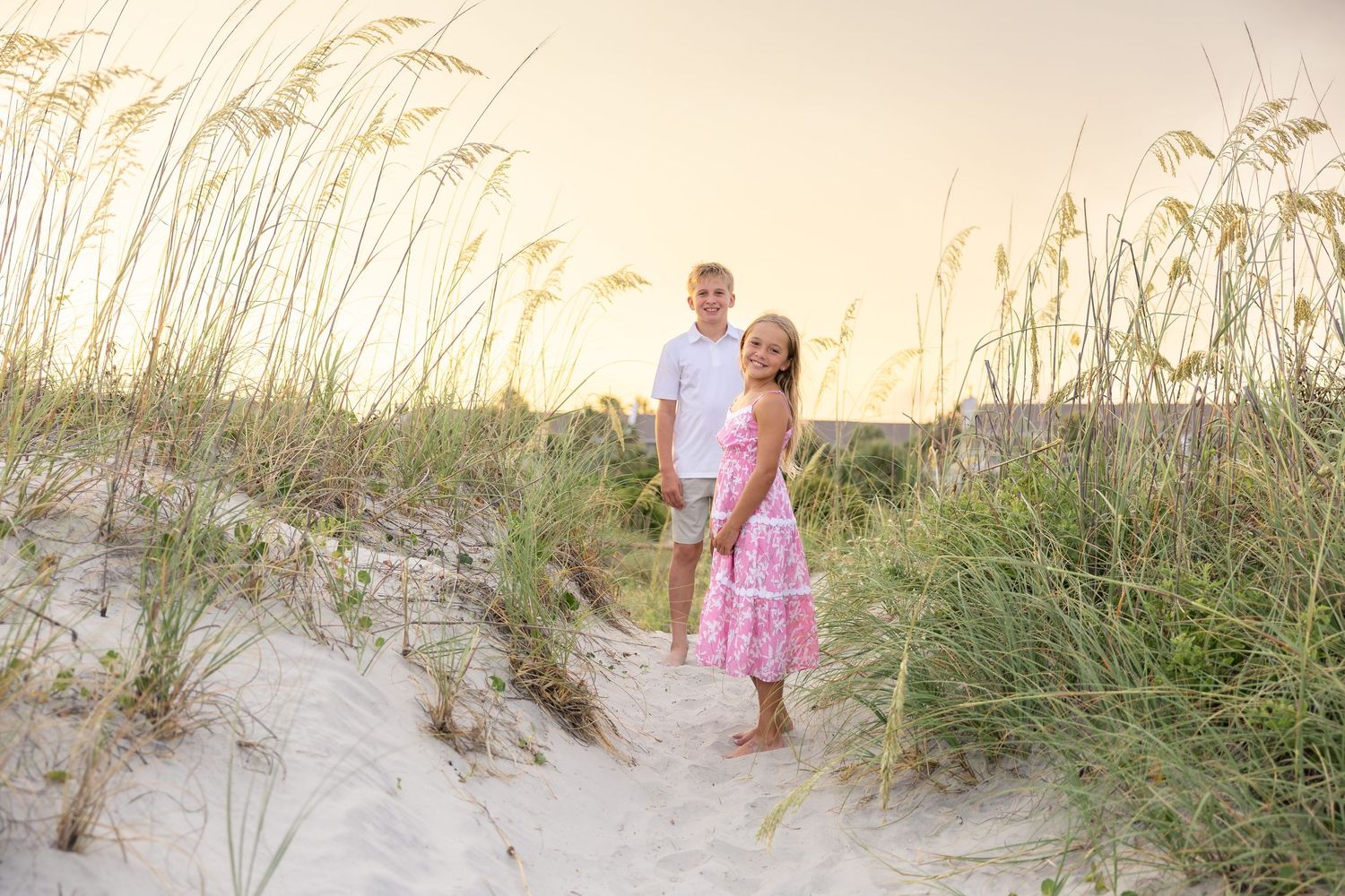 A couple poses on sandy beach dunes surrounded by sea oats at golden hour, woman in pink dress and man in white shirt.