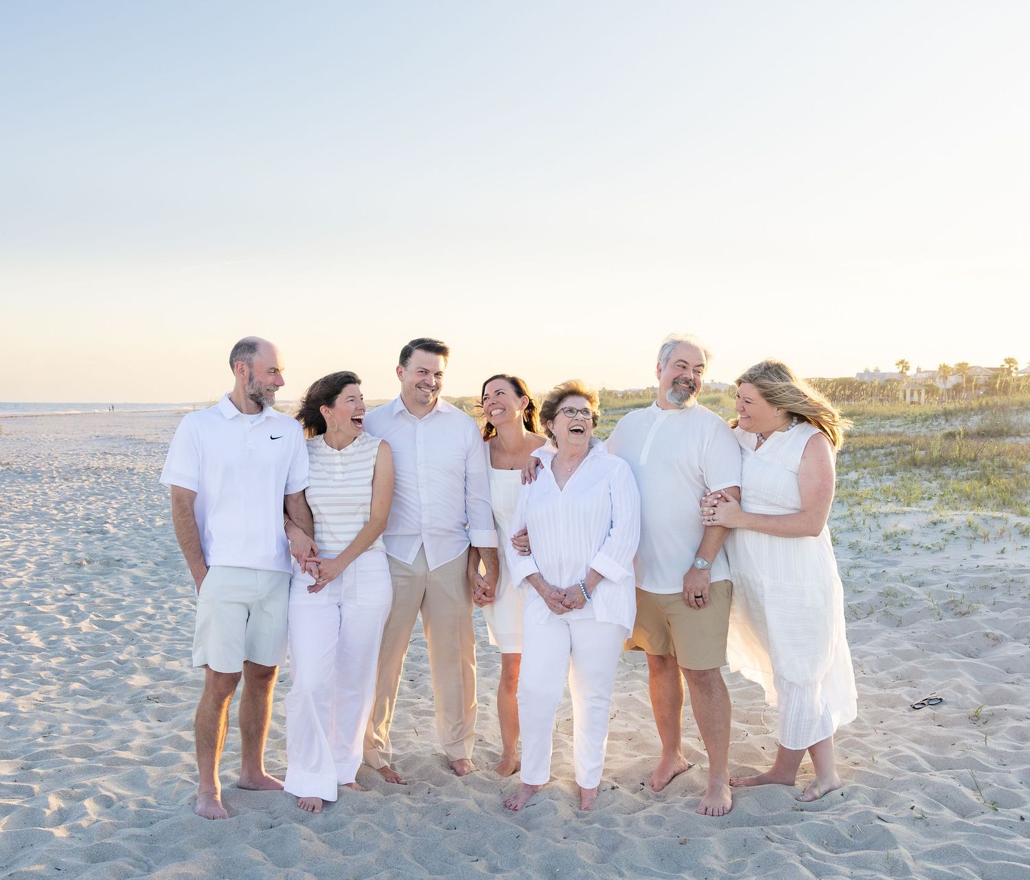 Family group in white outfits laughing together on a sunny beach at golden hour.