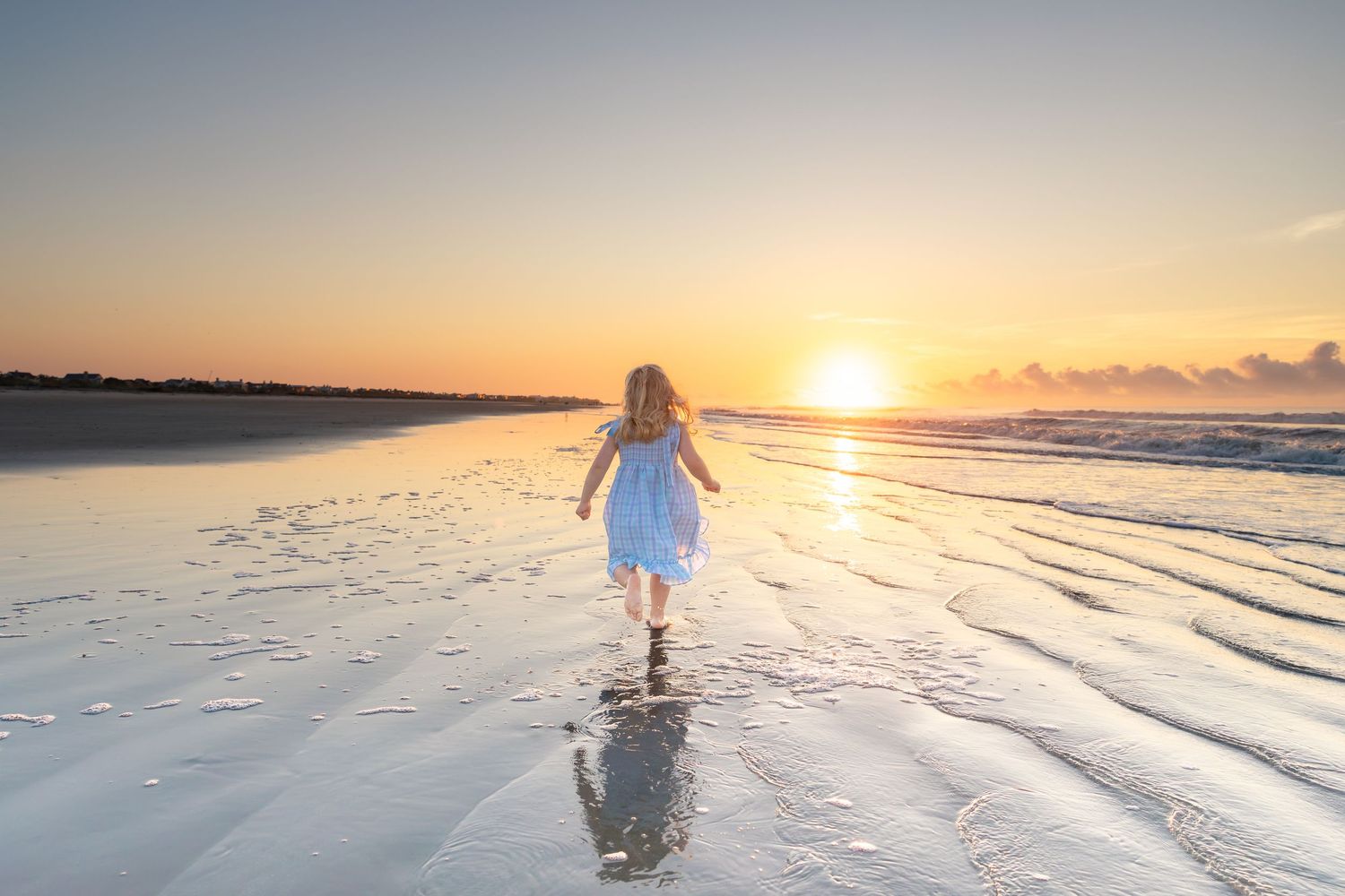 A young girl in a blue dress walks toward a golden sunset on a reflective wet beach.