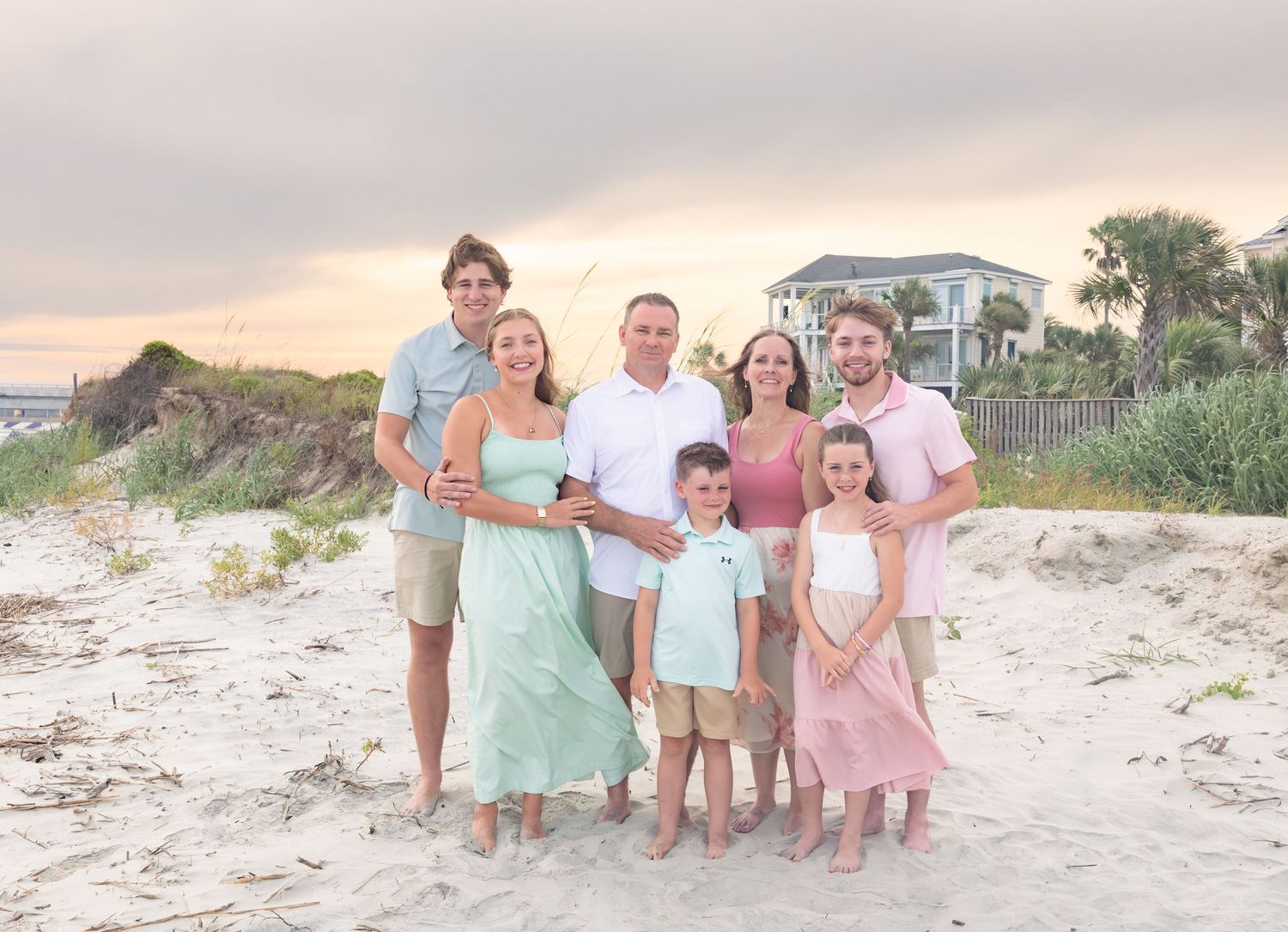 Family of seven posing together on a sandy beach at sunset with a coastal home and dunes in the background.