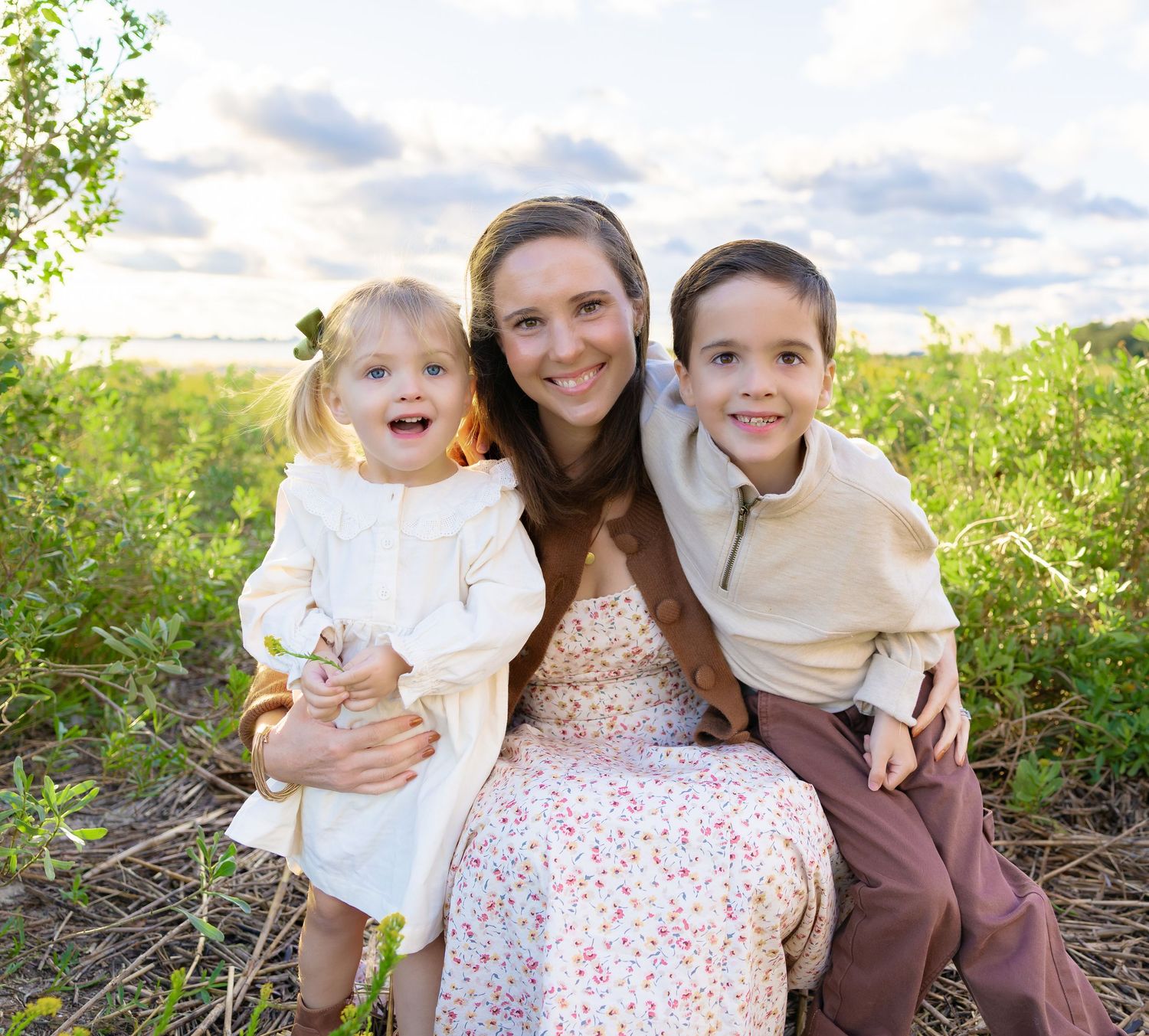 A smiling mother kneels in a grassy field at sunset with her young daughter and son hugging close, wearing coordinated neutral outfits.