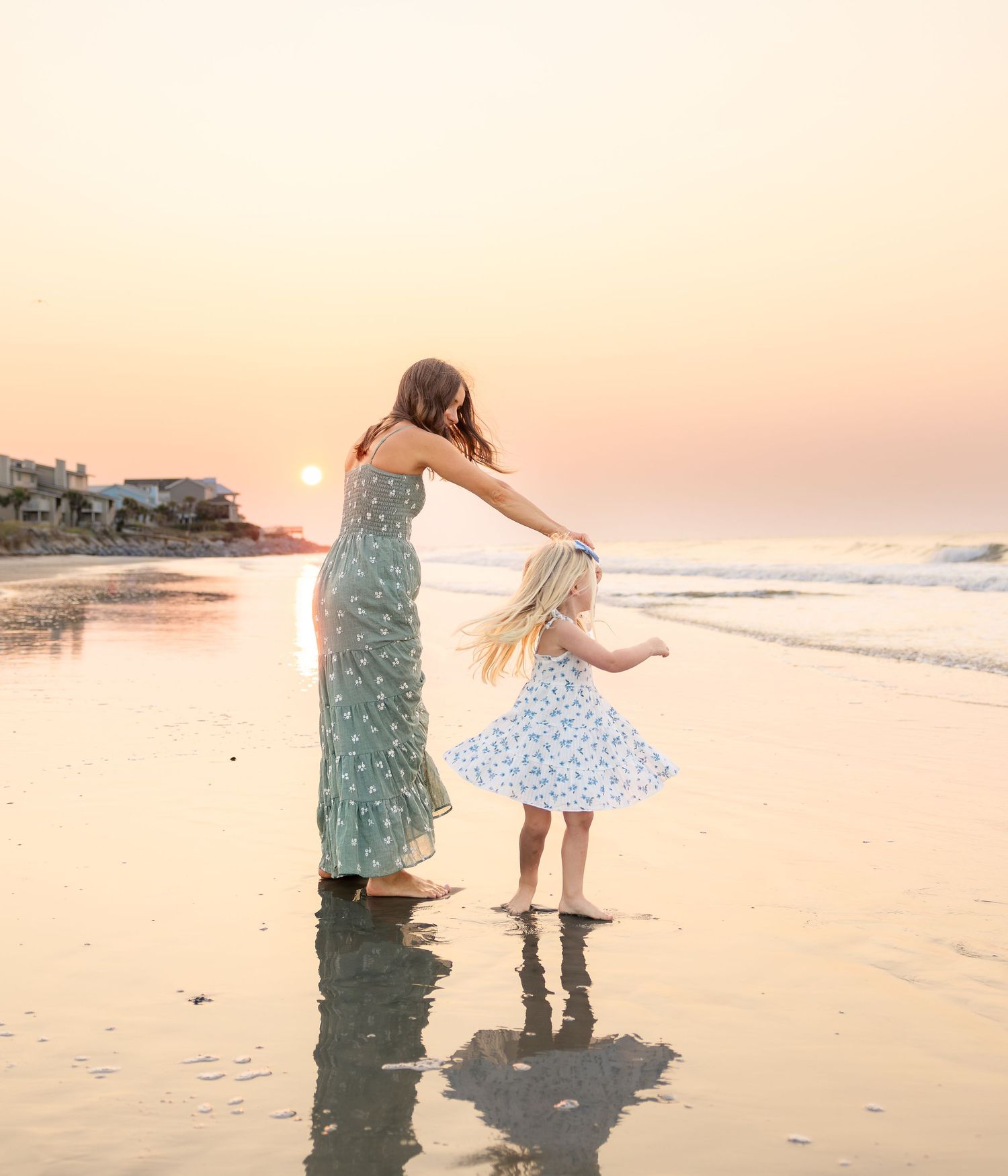 A mother and young daughter dance on a reflective beach at sunset, wearing floral dresses by the ocean waves.