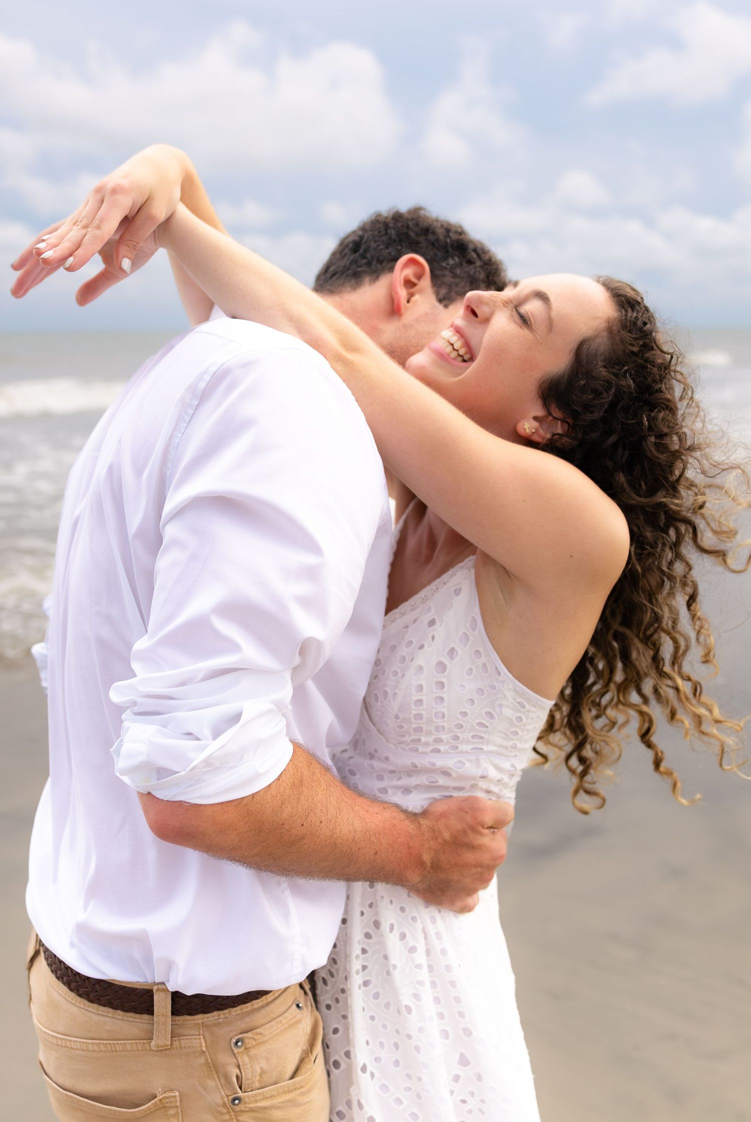 Happy couple dancing and embracing on a beach, wearing white outfits, with ocean waves in the background.