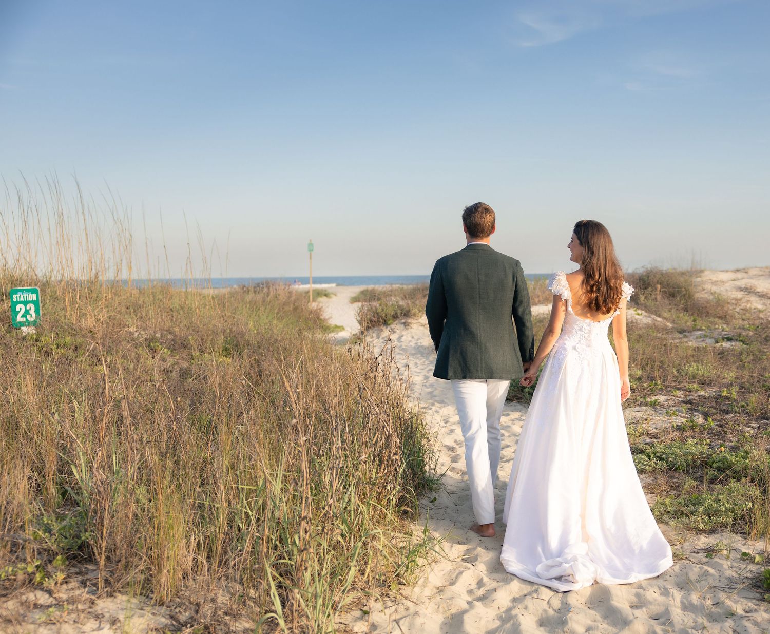 Bride and groom holding hands walking toward a sandy beach path surrounded by dunes and sea grass on their wedding day.