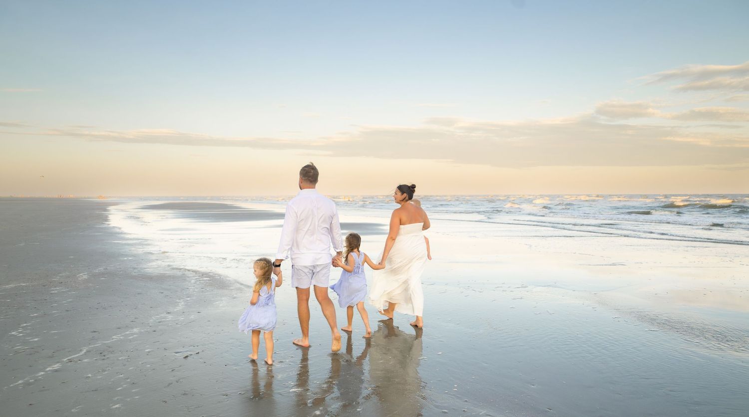 Family of four walking hand-in-hand along a serene beach at sunset, wearing white outfits with waves gently lapping the shore.