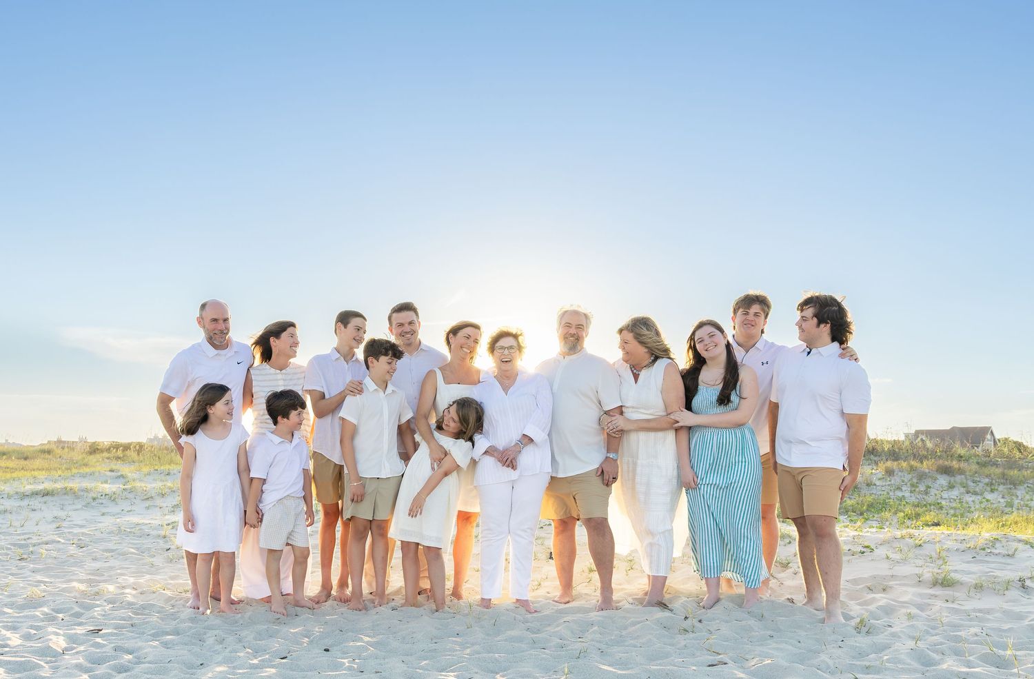 Large family posing together on a sunny beach, wearing white and neutral outfits, with blue sky backdrop.