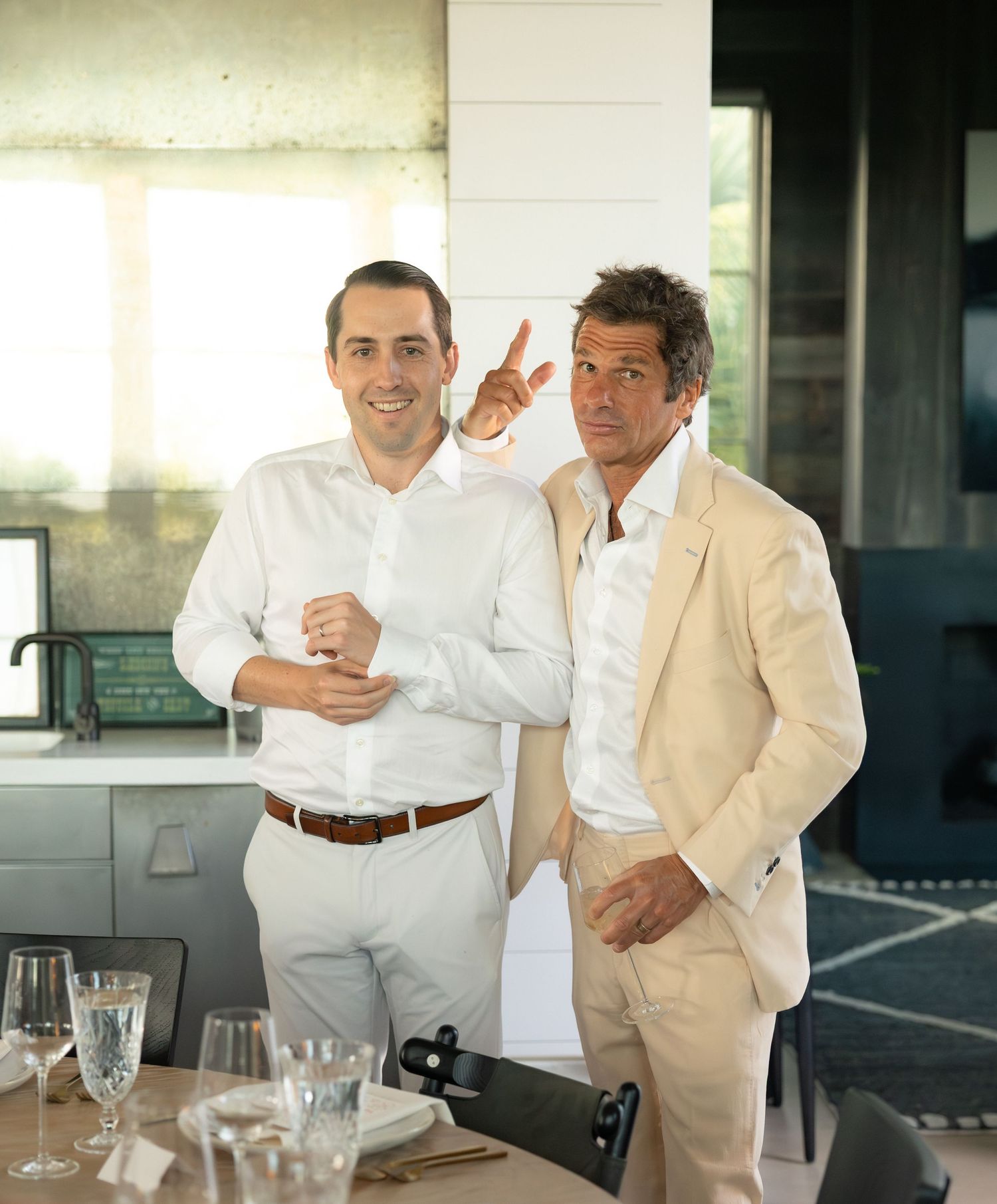 Two smiling men in white shirts pose indoors at wedding reception with glasses and table settings nearby.