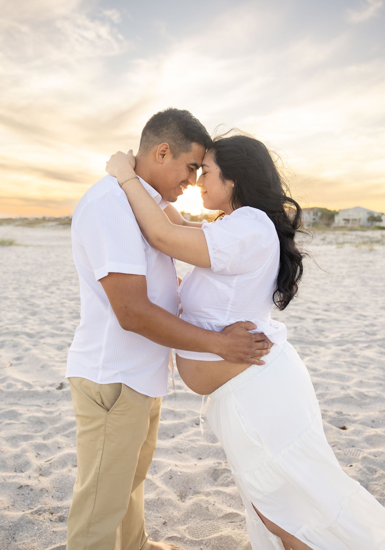 Couple in white embracing on sandy beach at sunset during maternity photoshoot, woman in flowing dress showing baby bump.