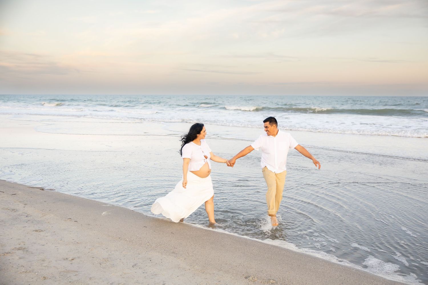 Pregnant couple holding hands and dancing on a sandy beach at sunset, woman in white dress, man in white shirt.