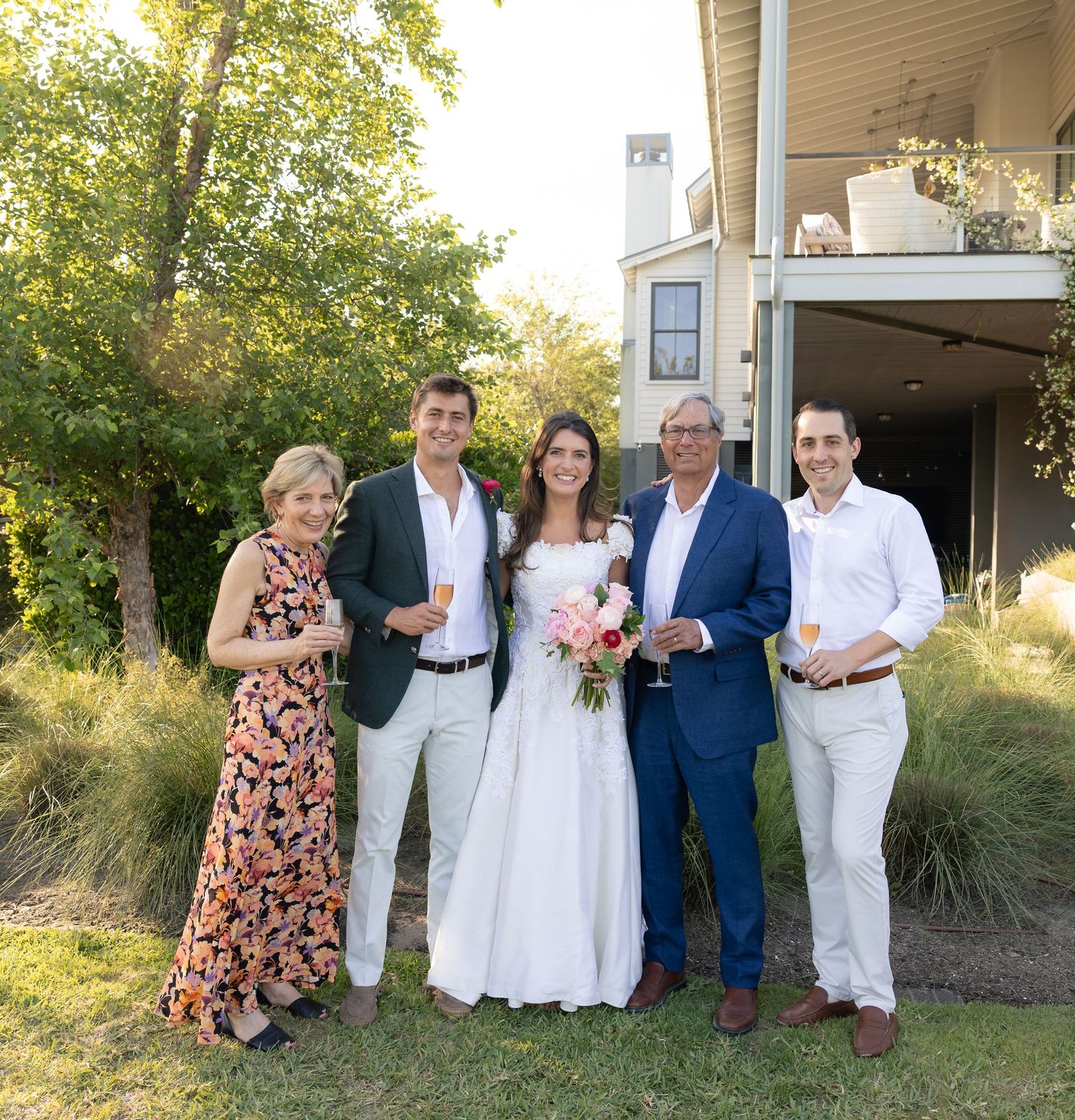 Wedding party of five smiling guests including bride with pink bouquet toasting with champagne in a garden setting.