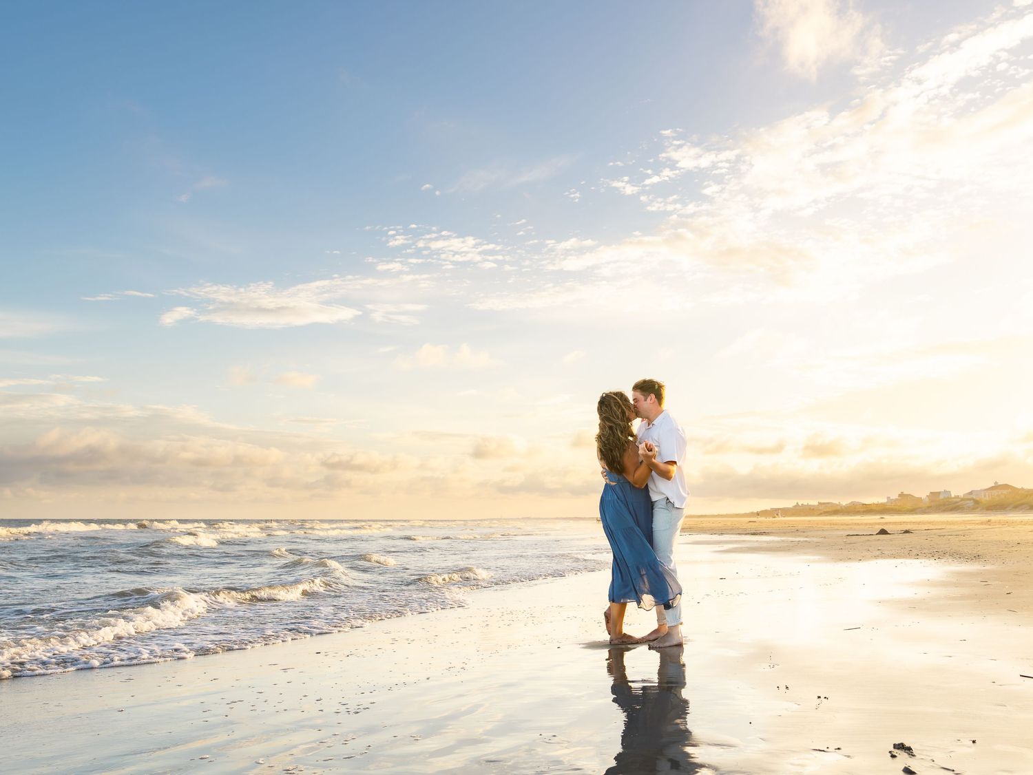 Couple embracing on a sandy beach at sunset with ocean waves and golden sky in the background.