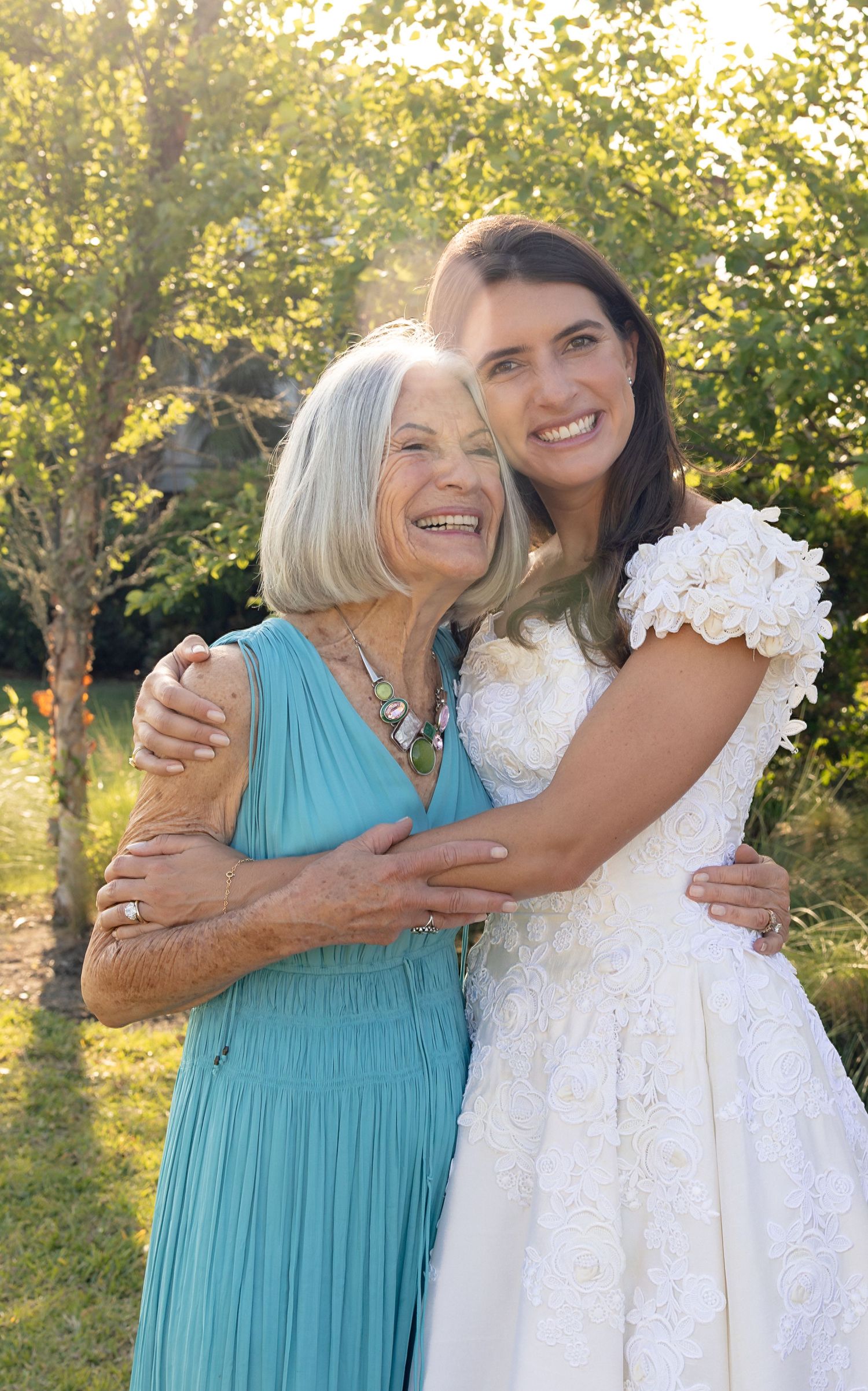 A smiling bride in a white lace wedding dress embraces an older woman in a teal dress outdoors among sunlit green trees.