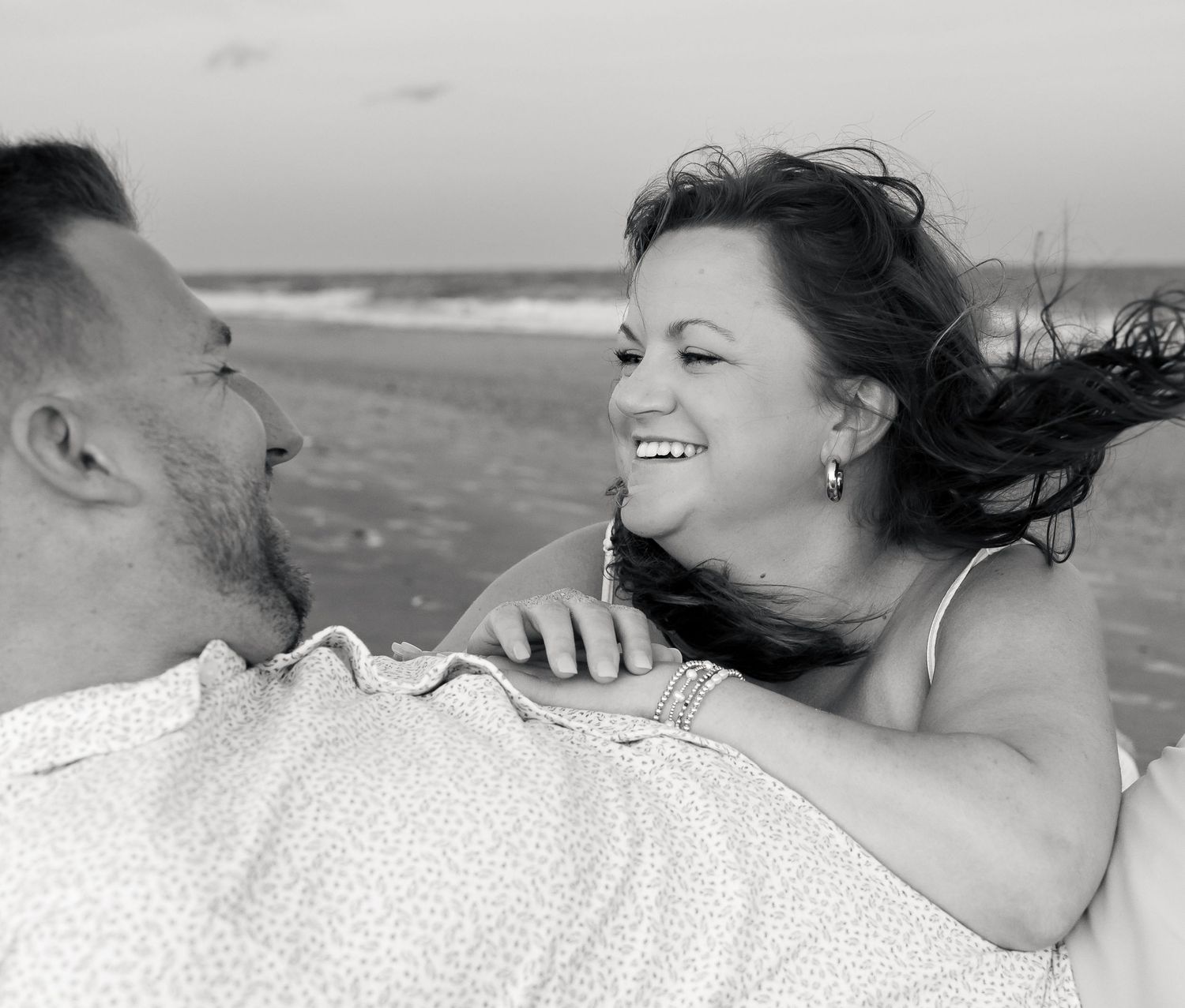 Black and white photo of a smiling woman with wind-blown hair laughing with a man on a beach at the ocean shore.