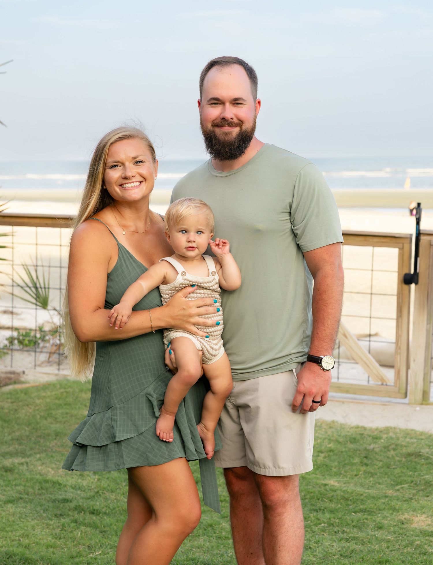 Smiling woman and bearded man in casual clothes holding a happy toddler on a grassy lawn with a beach and ocean behind them.