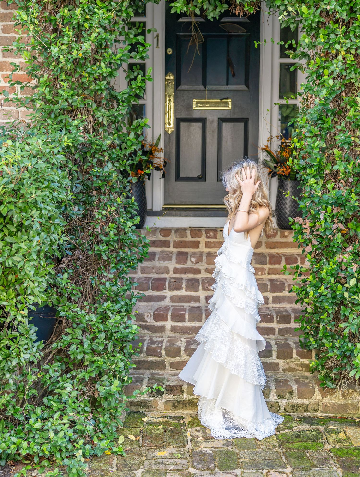 Girl in a white tiered ruffle gown poses with hands on head on brick steps before a black front door framed by lush green ivy.