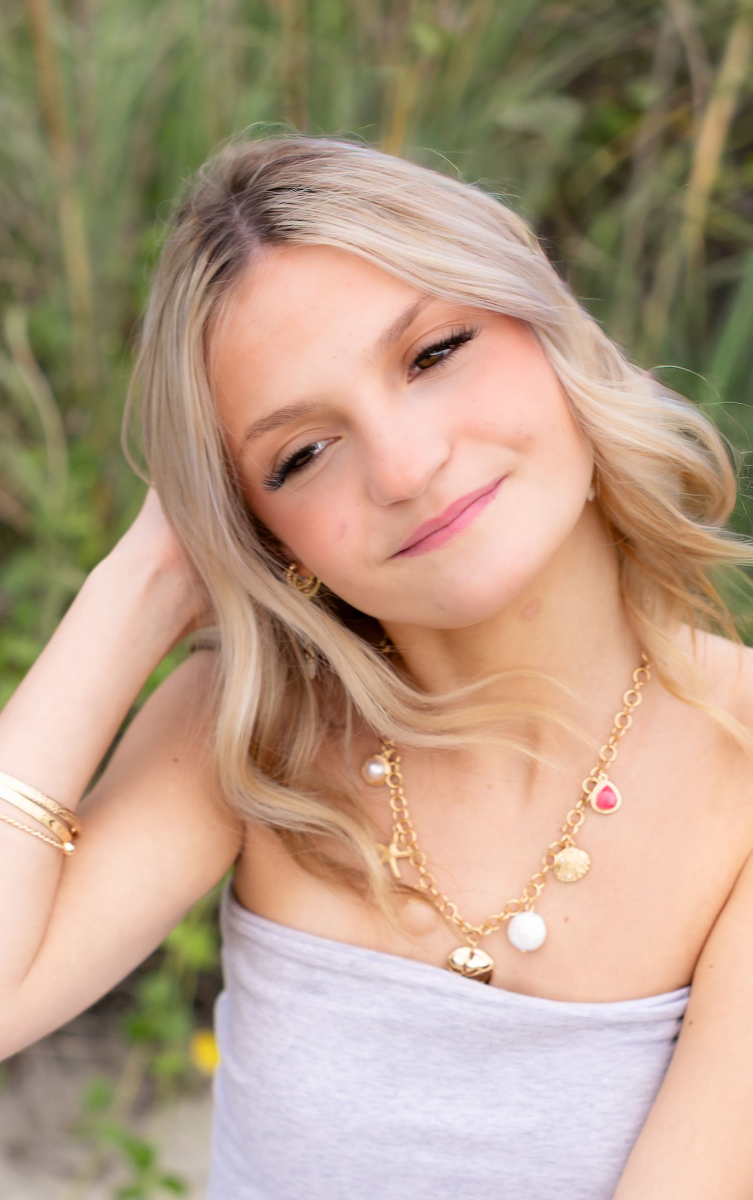 Smiling young woman with wavy blonde hair wearing a gray strapless top and gold charm necklace posing outdoors near tall green grass.