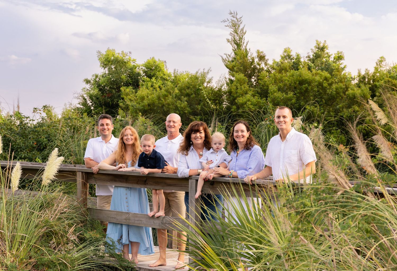 Large family group portrait outdoors on a wooden bridge surrounded by lush green trees and tall grass.