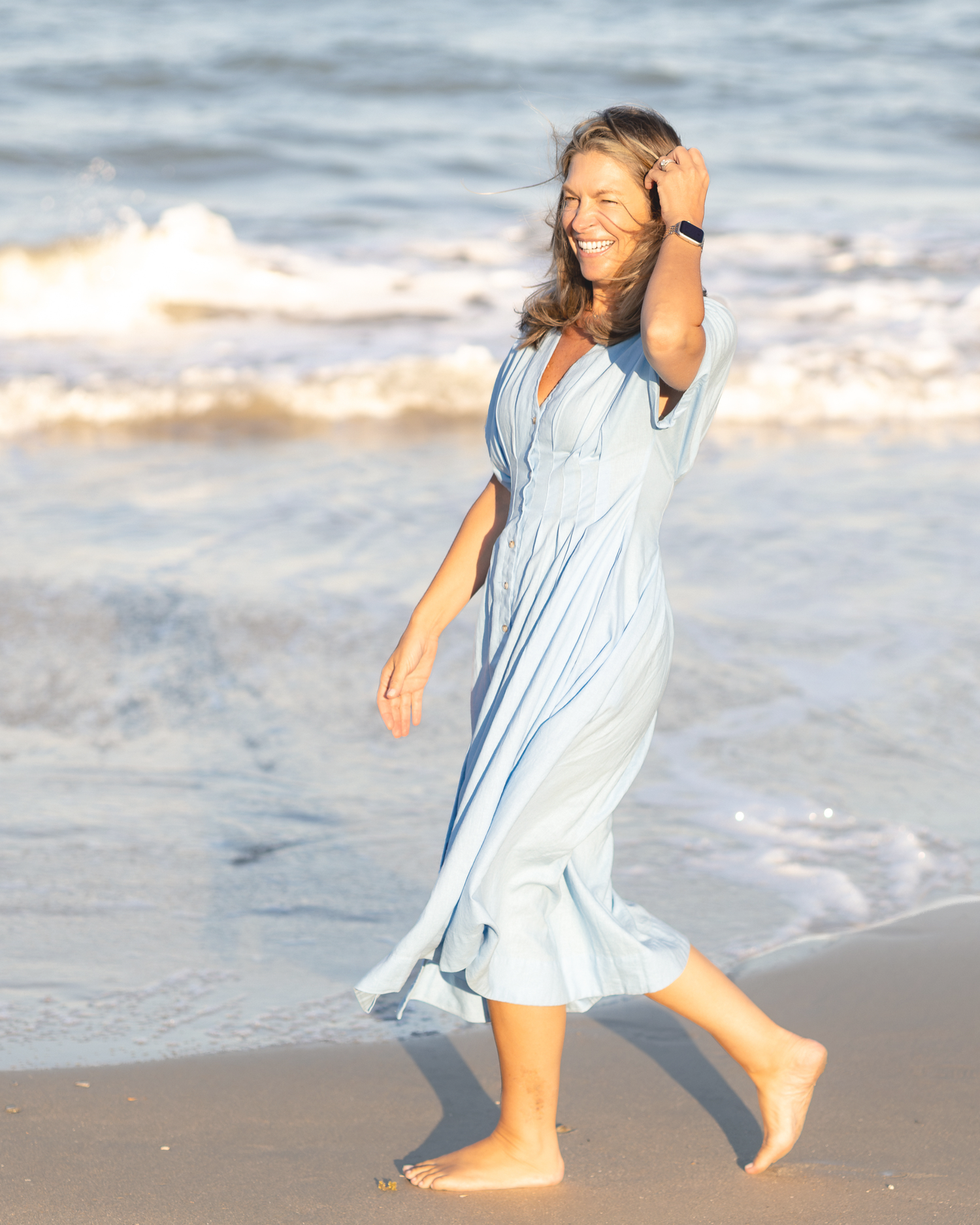 A smiling woman in a light blue maxi dress walks barefoot on a sunny beach with ocean waves behind her.