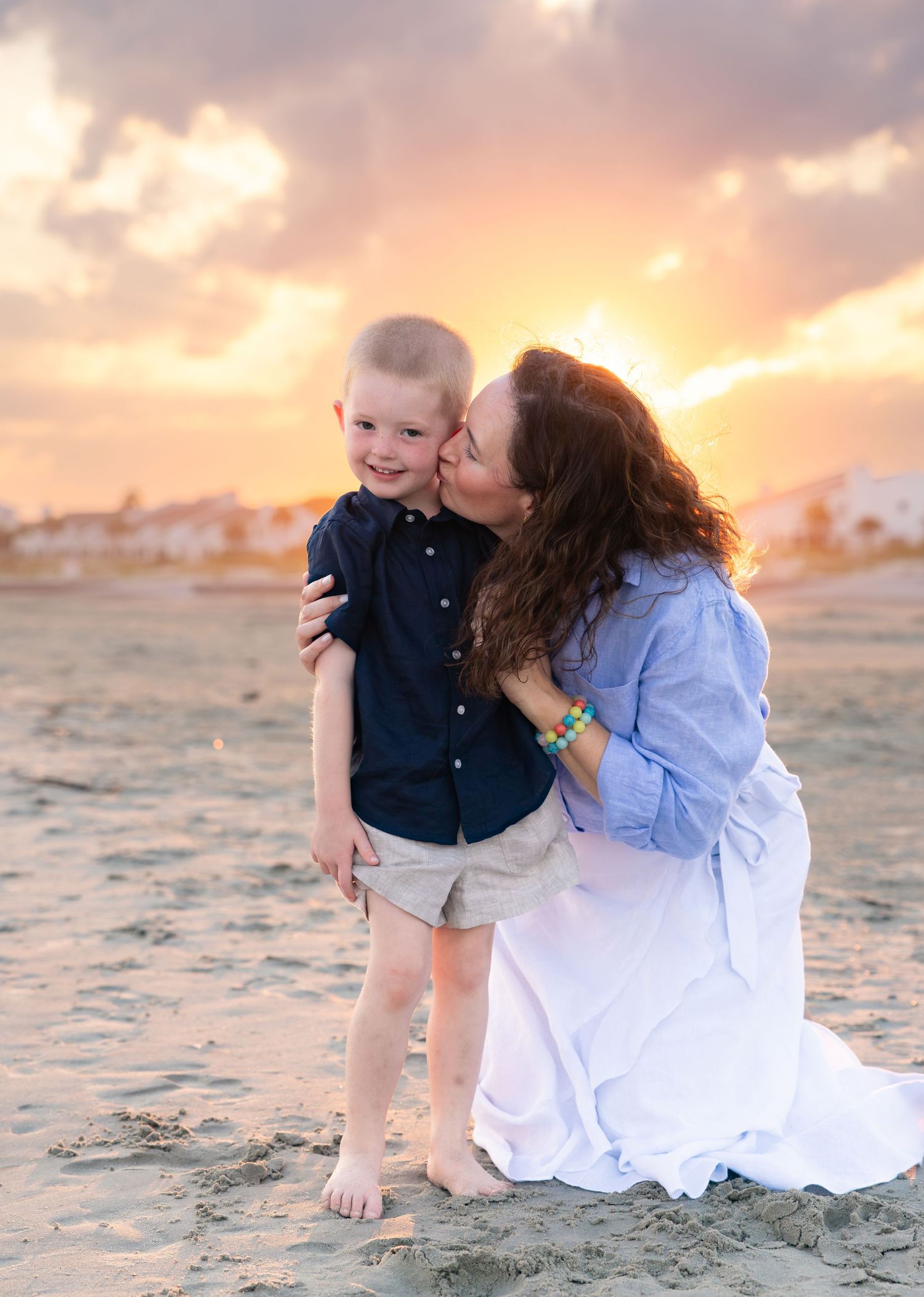 A mother kisses her young son on the cheek at sunset on a sandy beach, both dressed in casual coastal attire.
