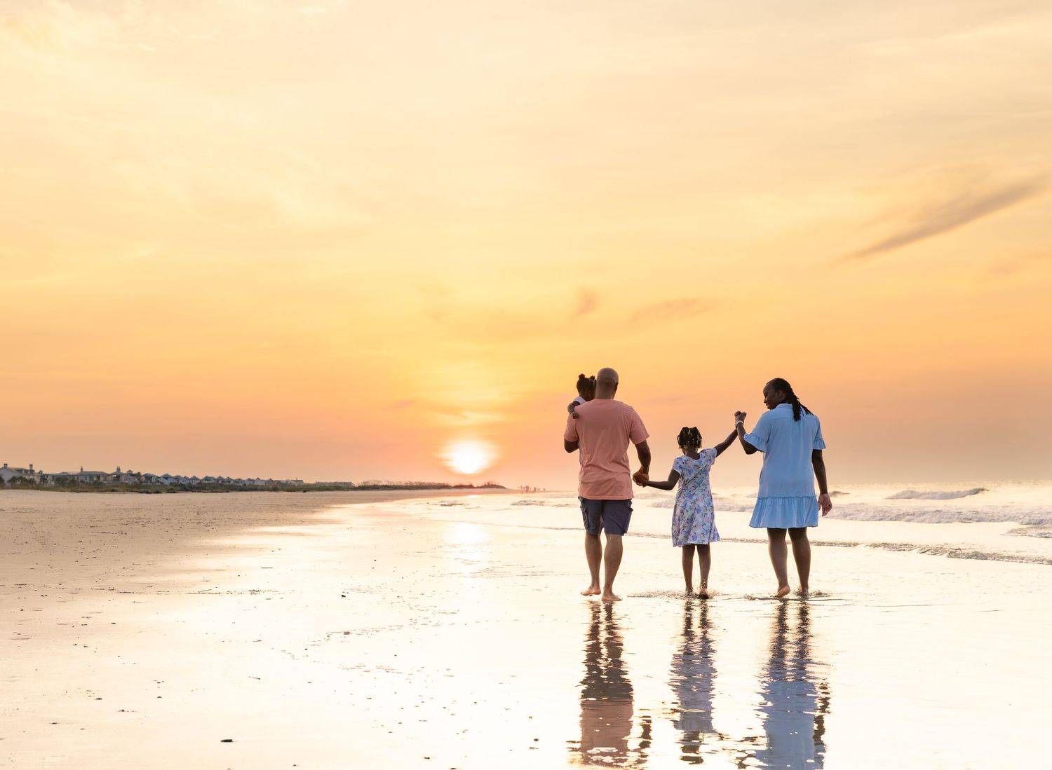 Family of four walking hand-in-hand on a beach at golden sunset, waves reflecting warm orange sky.
