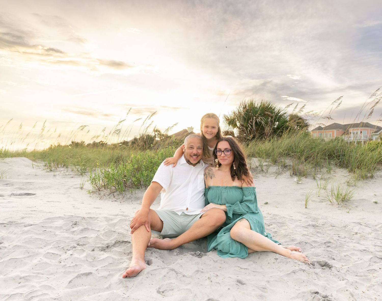 Family of three sitting on sandy beach dunes at sunset, wearing teal and white outfits, smiling warmly.
