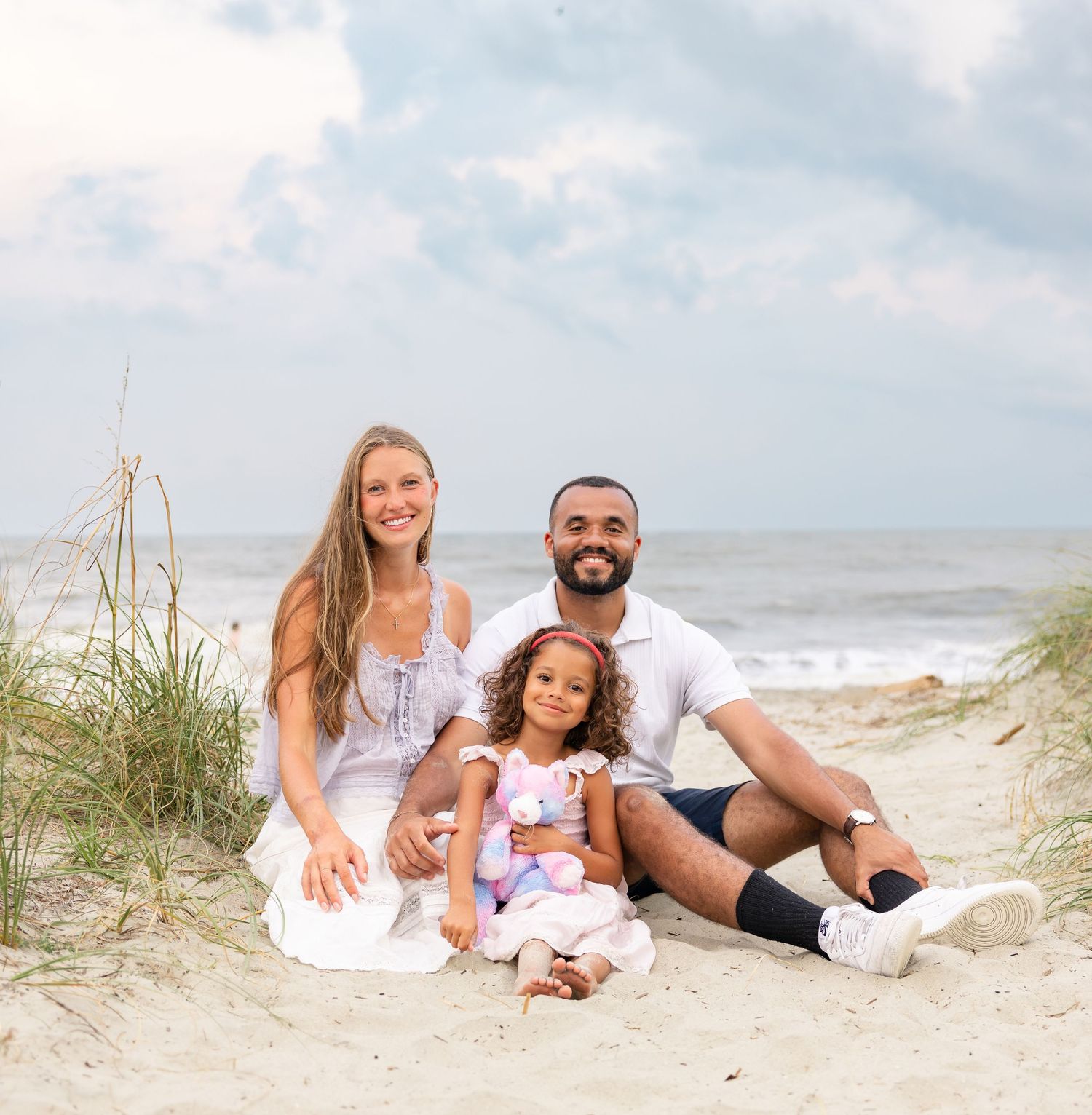 Family of three smiling together on a sandy beach, with ocean waves and cloudy sky in the background.